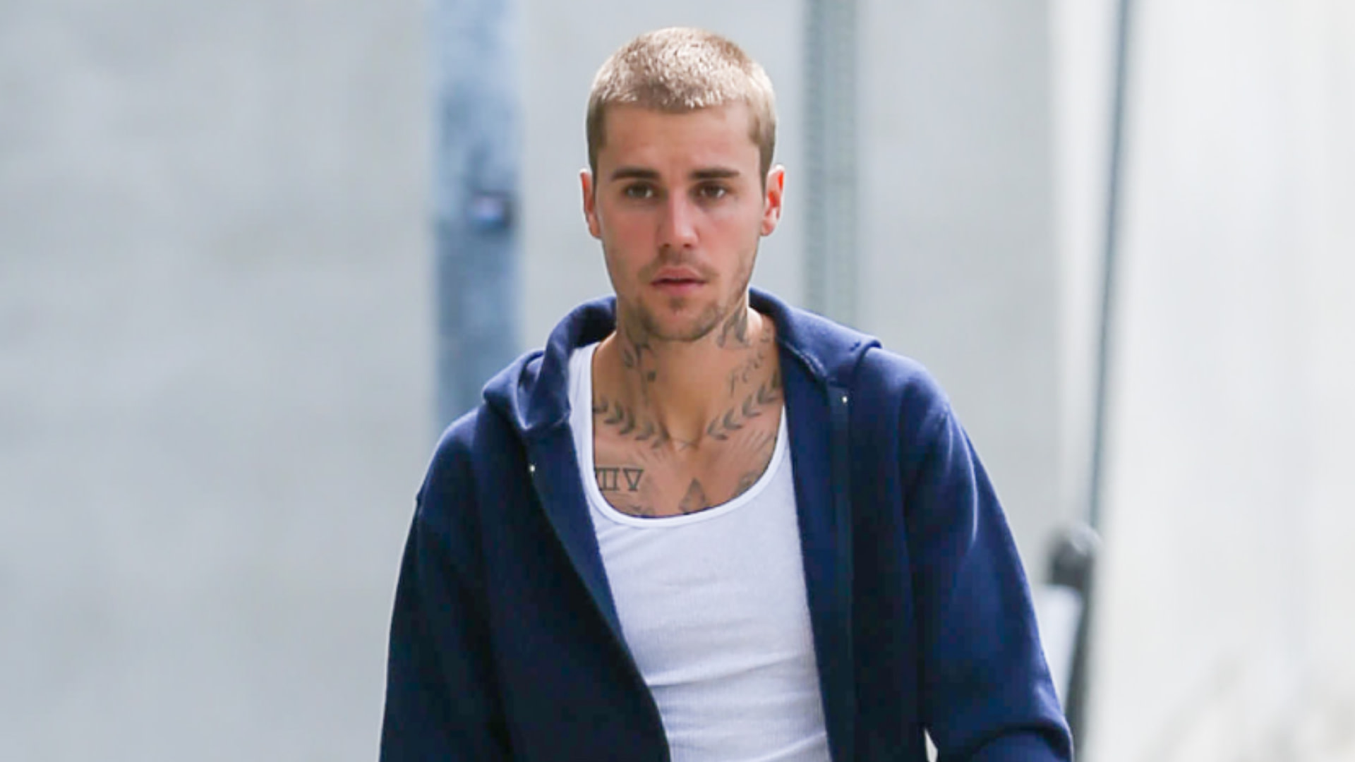 Justin Bieber with short hair and tattoos, wearing a navy hoodie and white tank top, walking outdoors.