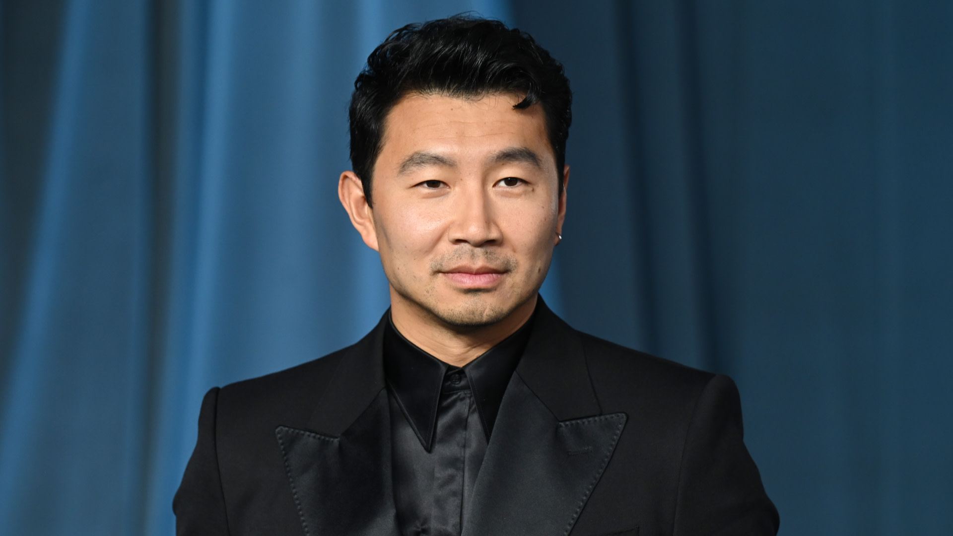Simu Liu in a black suit, standing against a blue curtain backdrop, looking confident.