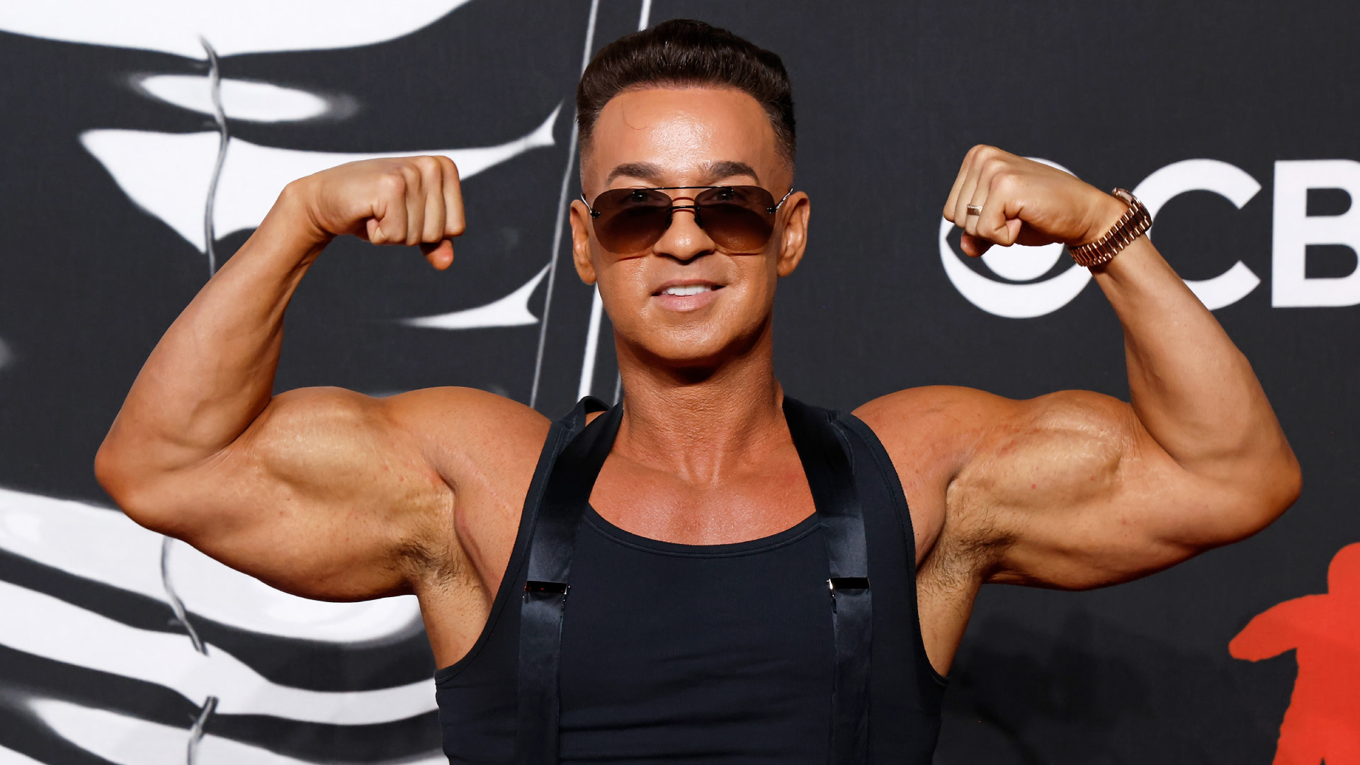 Mike "The Situation" Sorrentino wearing sunglasses flexes his biceps on a red carpet, dressed in a black tank top with a patterned background.