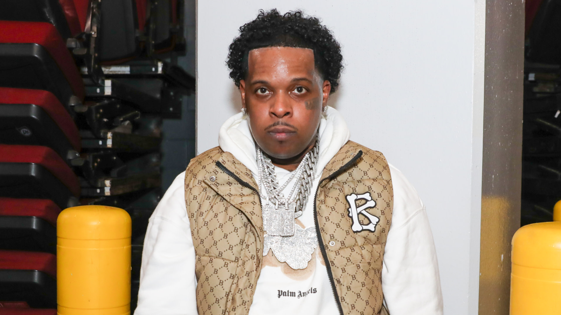 Finesse2tymes wearing a Gucci jacket and a Palm Angels hoodie with layered chains, standing indoors near yellow bollards.