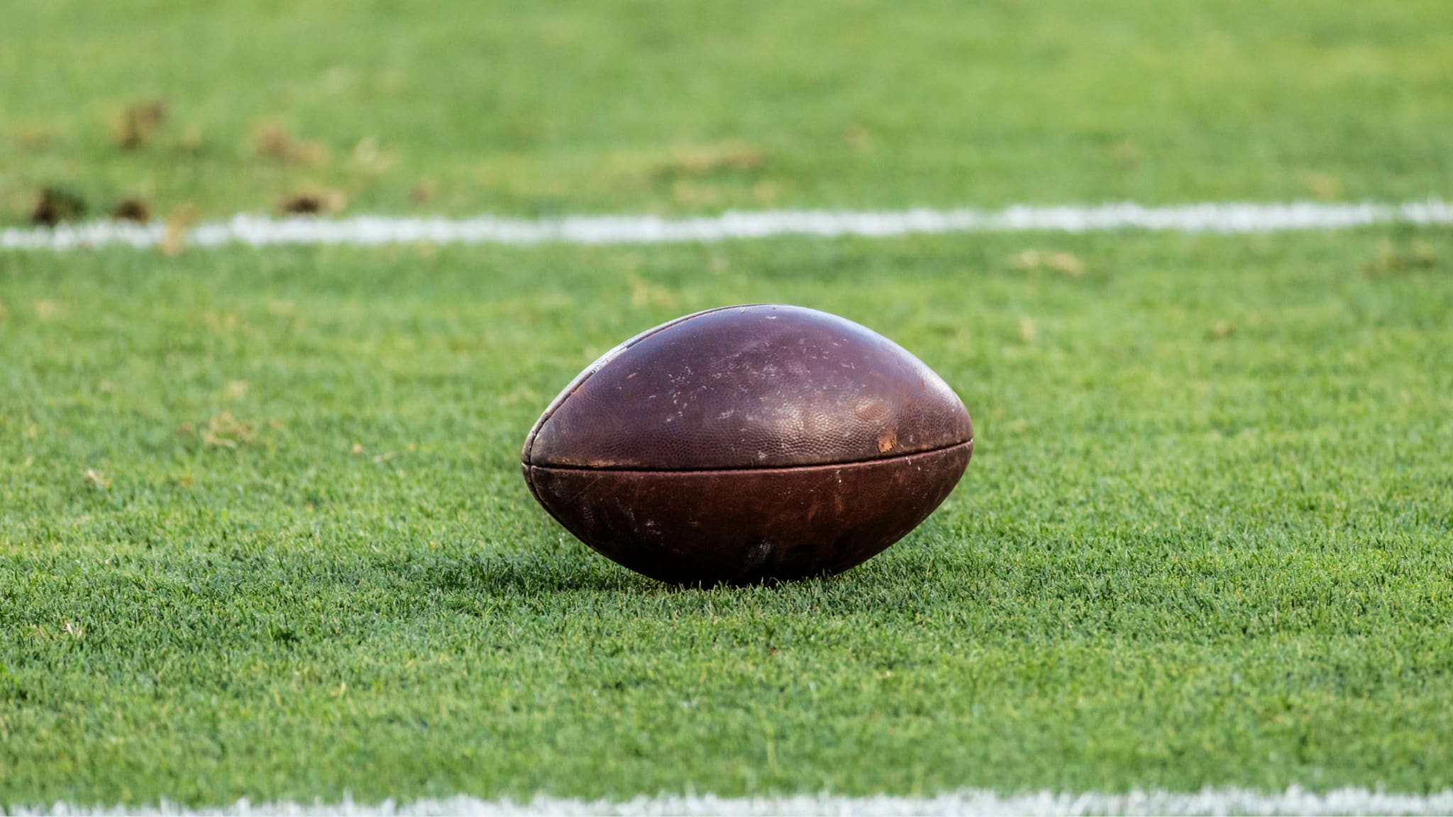 A vintage leather football rests on a grassy field, with white yard lines visible in the background.