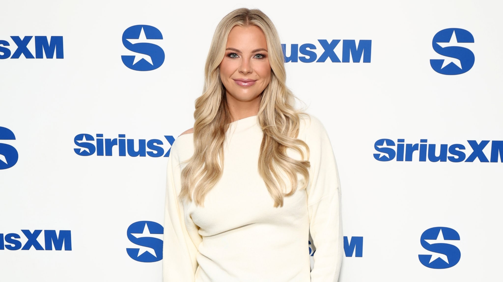 A woman with long blonde hair in a white outfit stands in front of a SiriusXM backdrop, smiling.