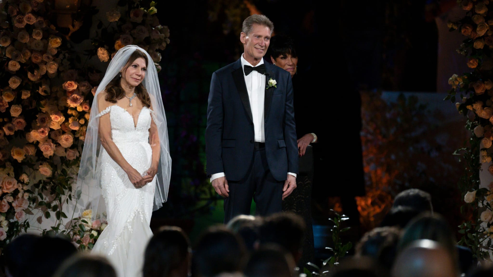 A bride and groom stand at the altar during a wedding ceremony, surrounded by flowers and guests.