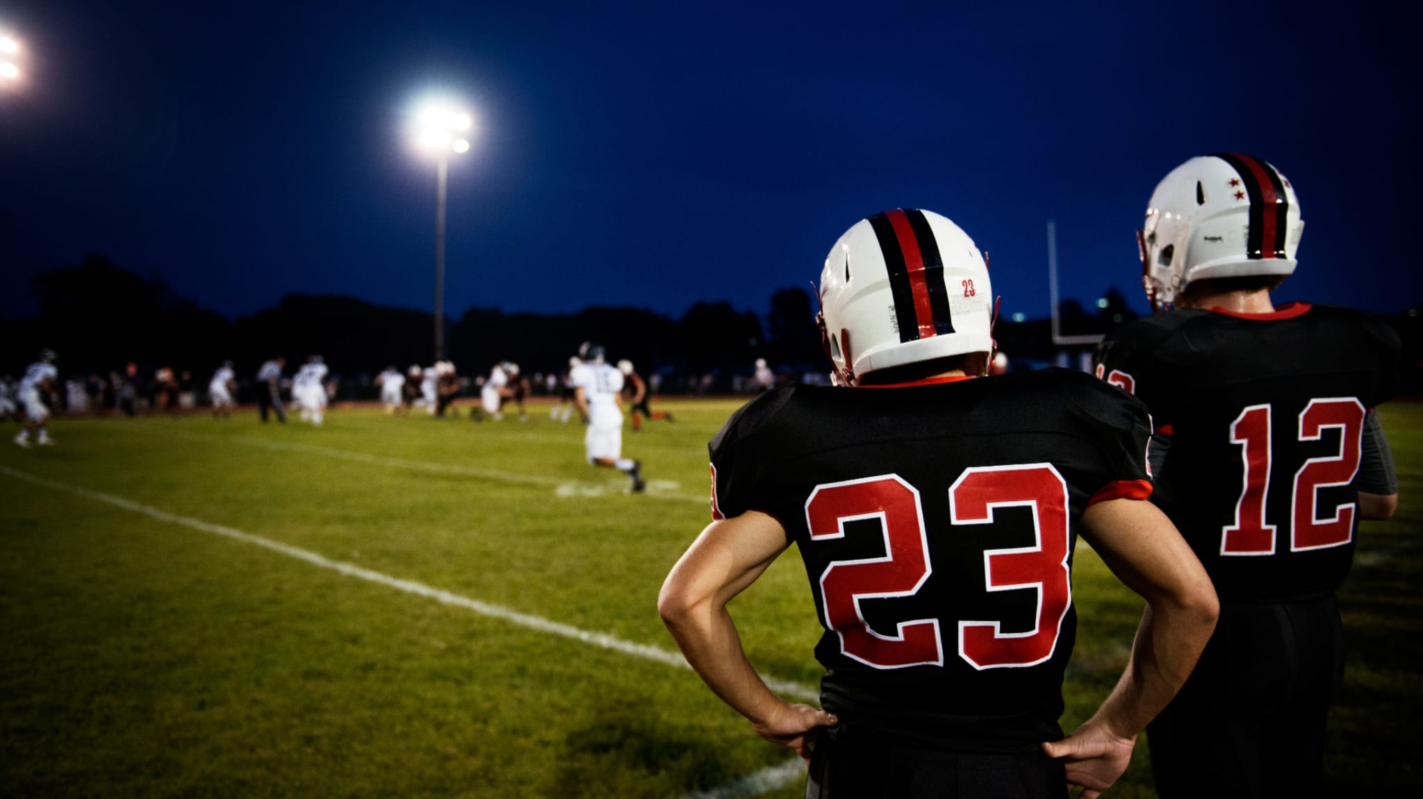 Two football players in black uniforms with numbers 23 and 12 watch a night game under bright stadium lights.