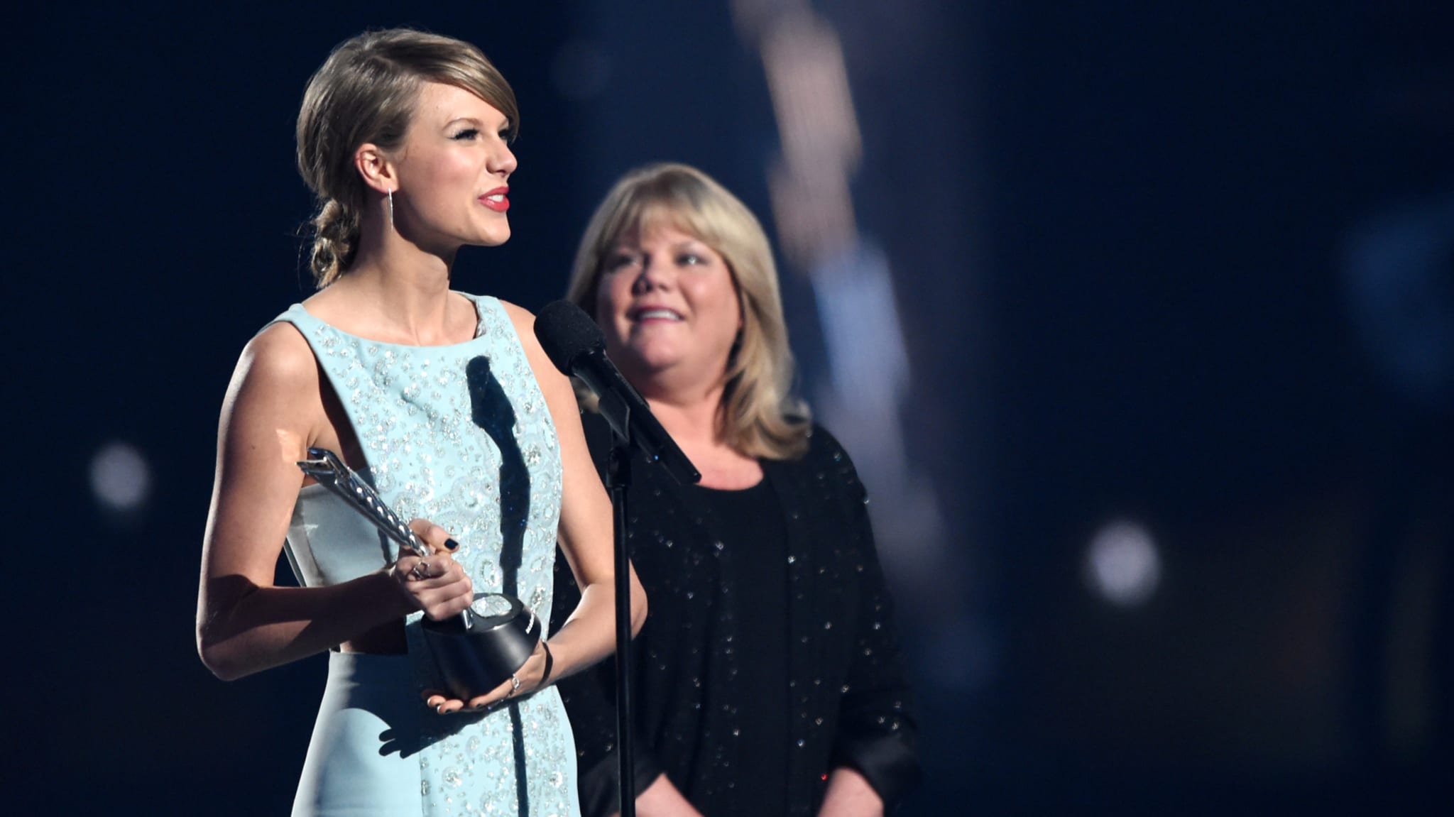 Taylor Swift holding an award, wearing a light blue dress, speaks into a microphone. Another woman stands beside her, smiling.