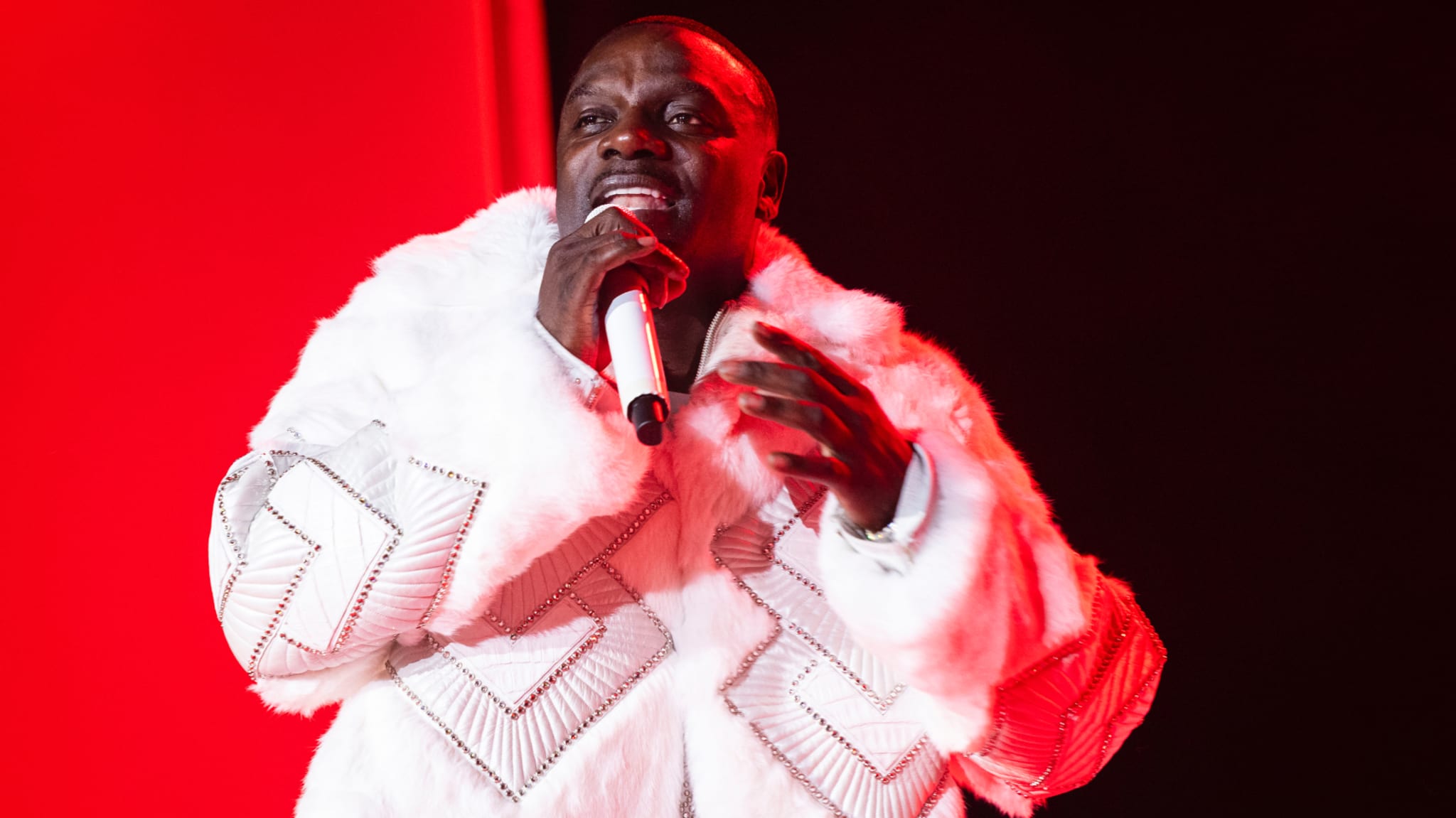 Akon performing on stage in a white fur coat, holding a microphone against a red background.