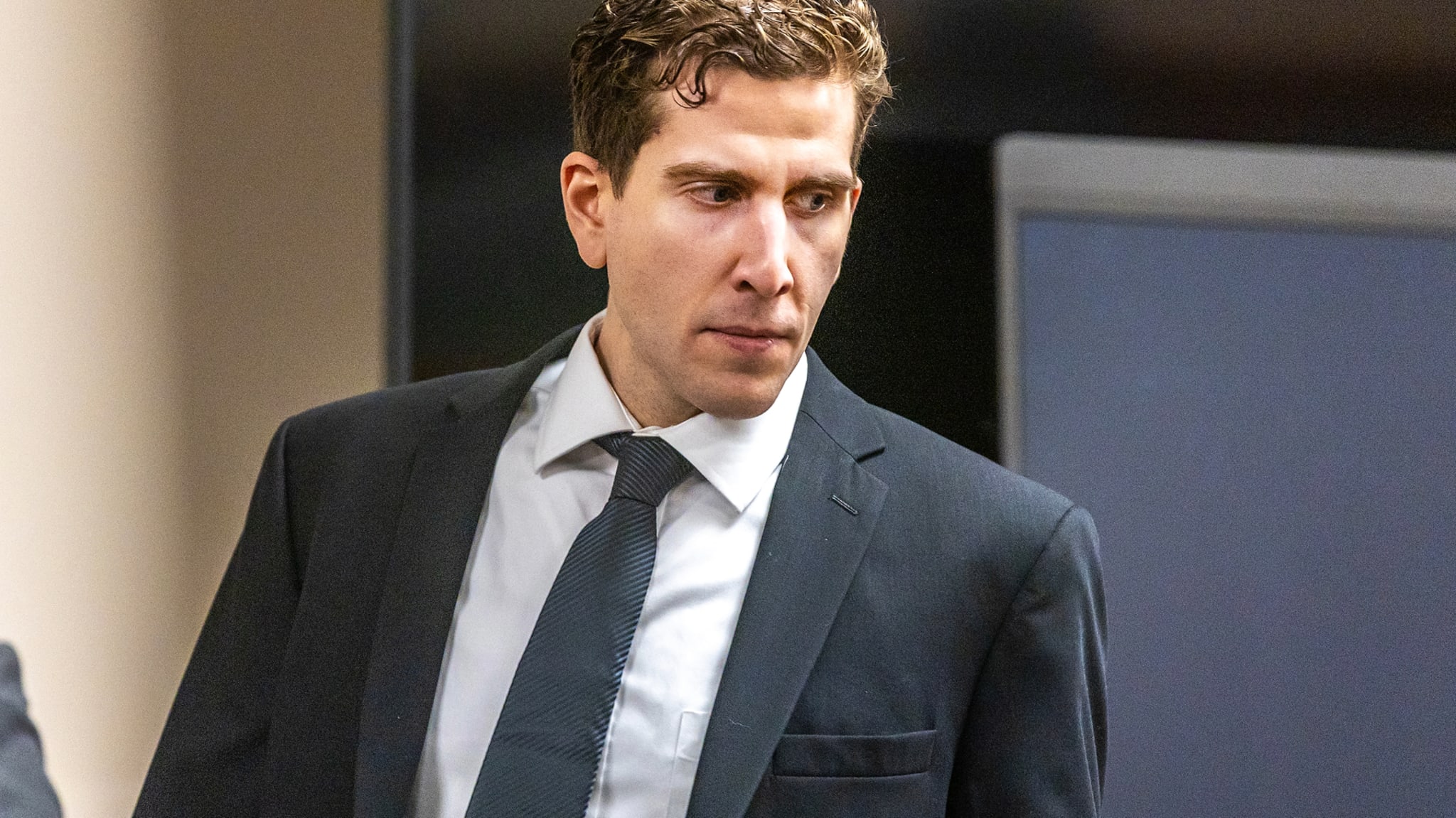 A man in a suit and tie stands indoors, looking to the side, with a serious expression.