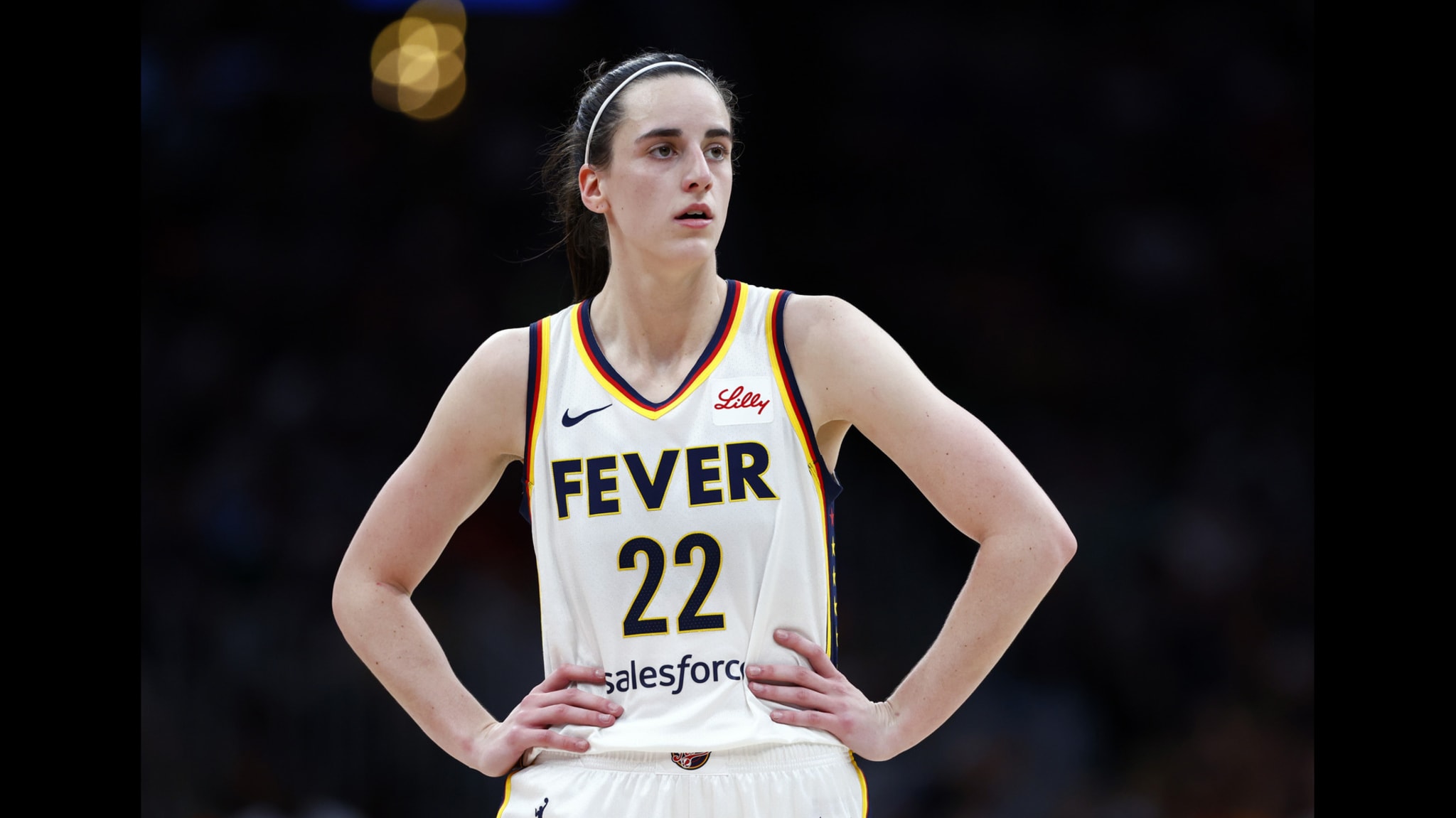Caitlin Clark wearing a "Fever" jersey with the number 22 stands on the court with hands on hips, looking focused.