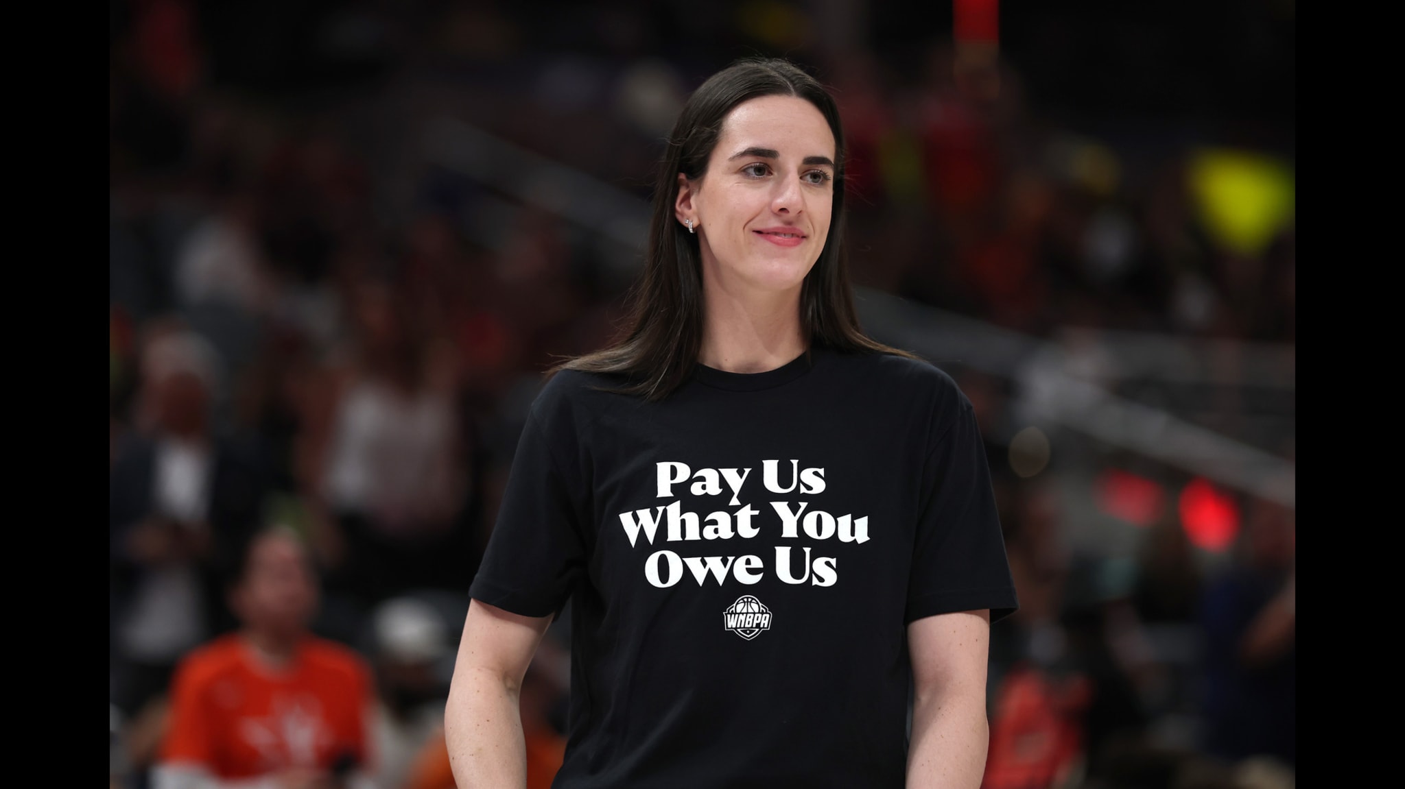 Caitlin Clark in a black T-shirt with the text "Pay Us What You Owe Us" stands in a sports arena.