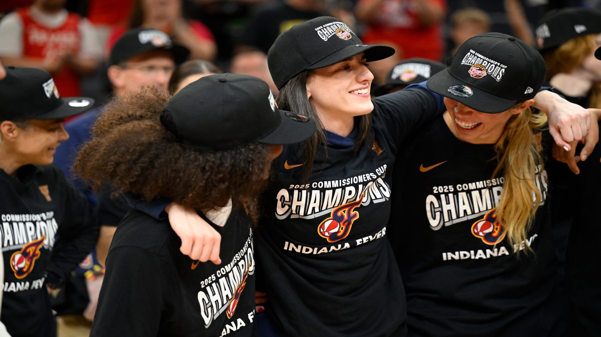 A group of Indiana Fever players celebrating, wearing "2025 Commissioner's Cup Champions" shirts and caps, smiling and embracing.