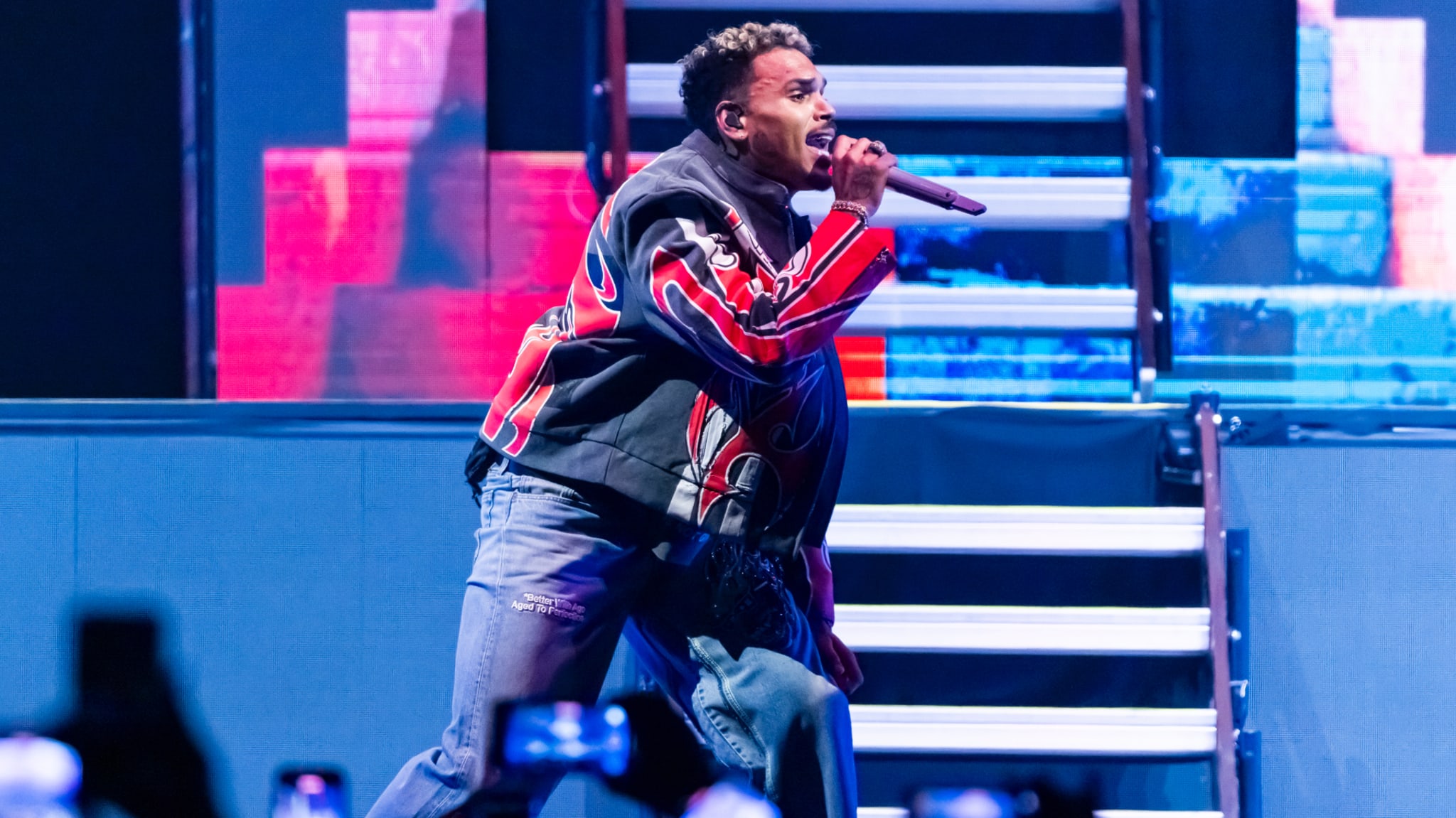 Chris Brown in a colorful jacket performs energetically on stage, holding a microphone, with vibrant lighting and steps in the background.