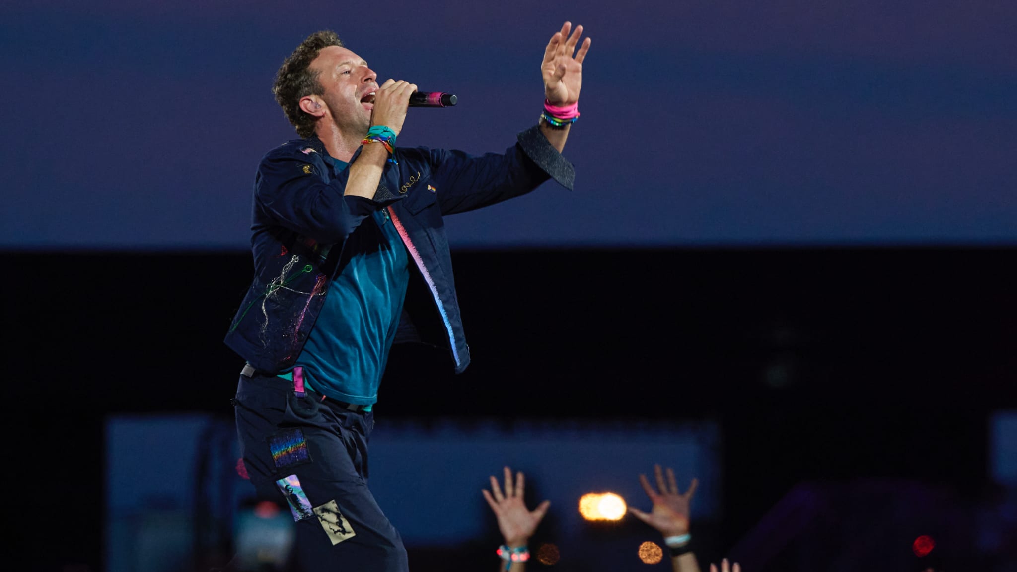Chris Martin performing on stage, wearing a colorful outfit, passionately singing into a microphone with raised hands in the audience.