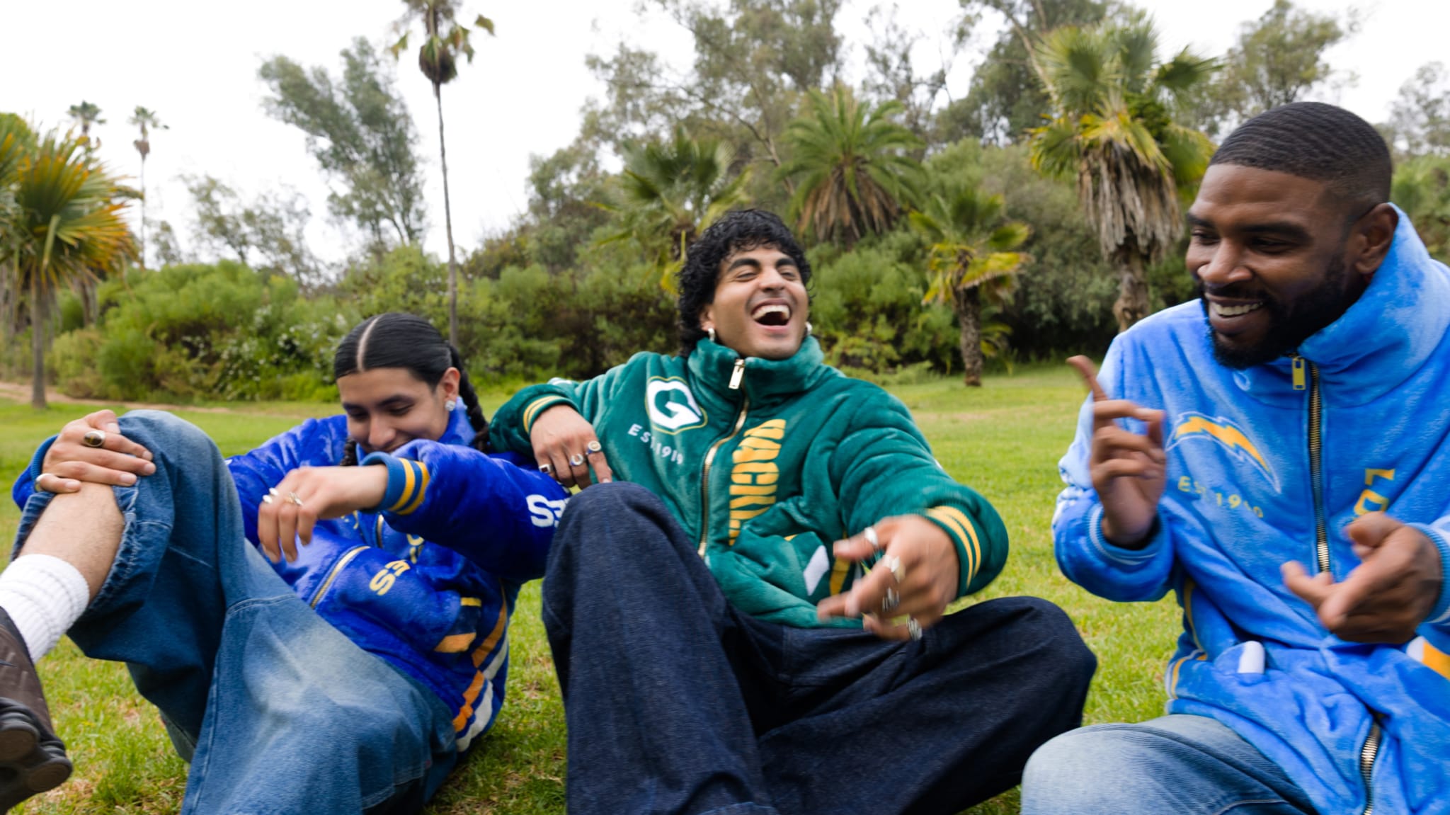 Three people sitting on grass, laughing and wearing colorful jackets. Palm trees and greenery in the background.