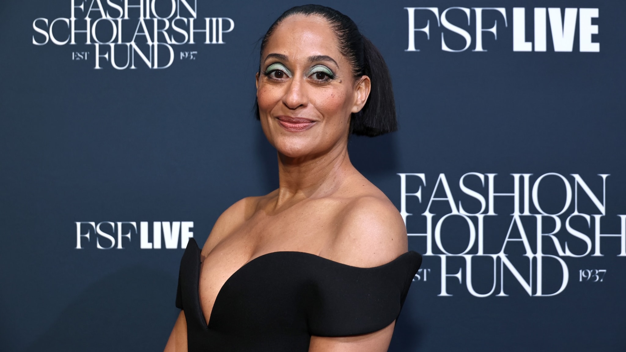 Tracee Ellis Ross at the Fashion Scholarship Fund event, wearing a black off-shoulder dress, with bold eye makeup and a sleek hairstyle.