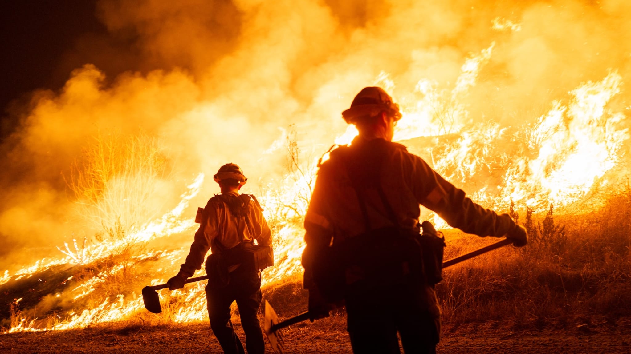 Firefighters battling a large wildfire at night, with flames and smoke in the background.