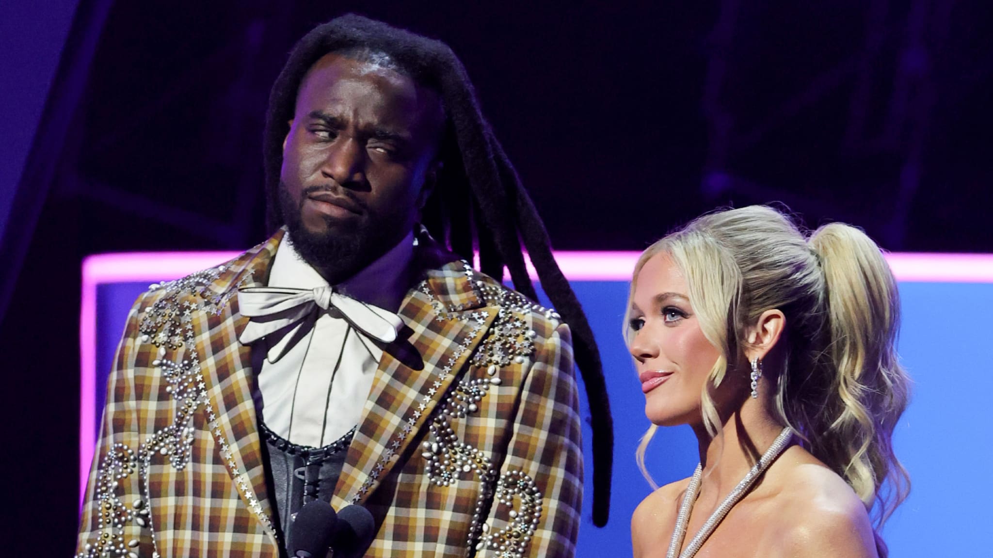 Two people on stage, Shaboozey with long dreadlocks and a plaid suit, Megan Moroney with blonde hair in a ponytail, wearing a strapless dress.