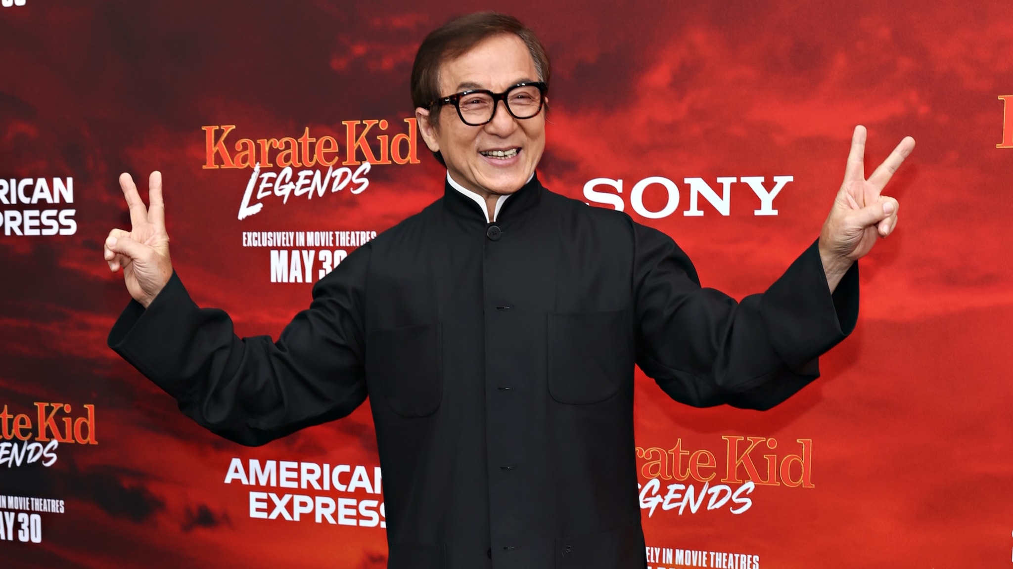 Jackie Chan at the "Karate Kid Legends" event, smiling and flashing peace signs, wearing a black outfit.