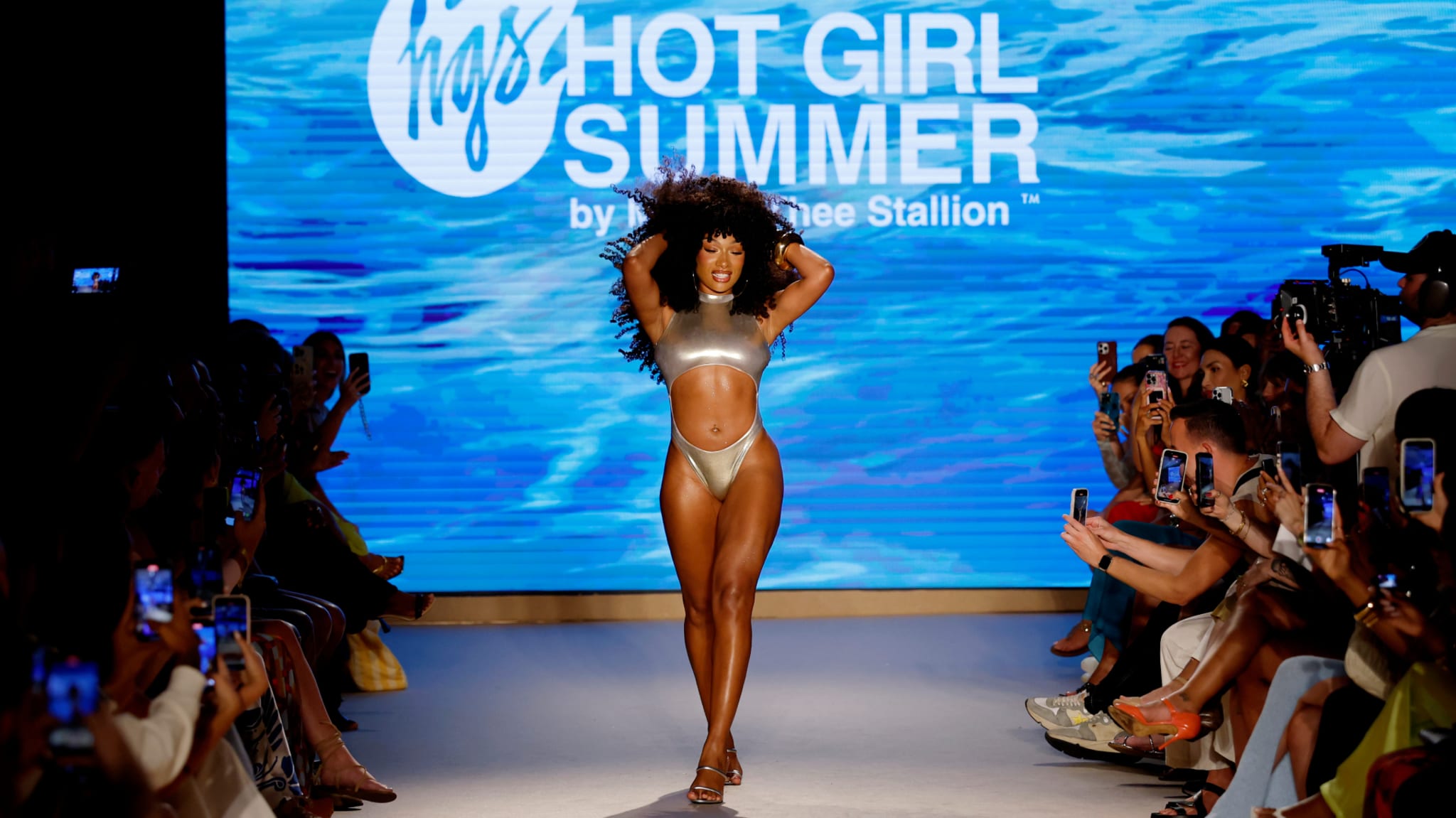 Megan Thee Stallion walks the runway in a silver bikini at a fashion show, with a "Hot Girl Summer" backdrop and audience capturing photos.