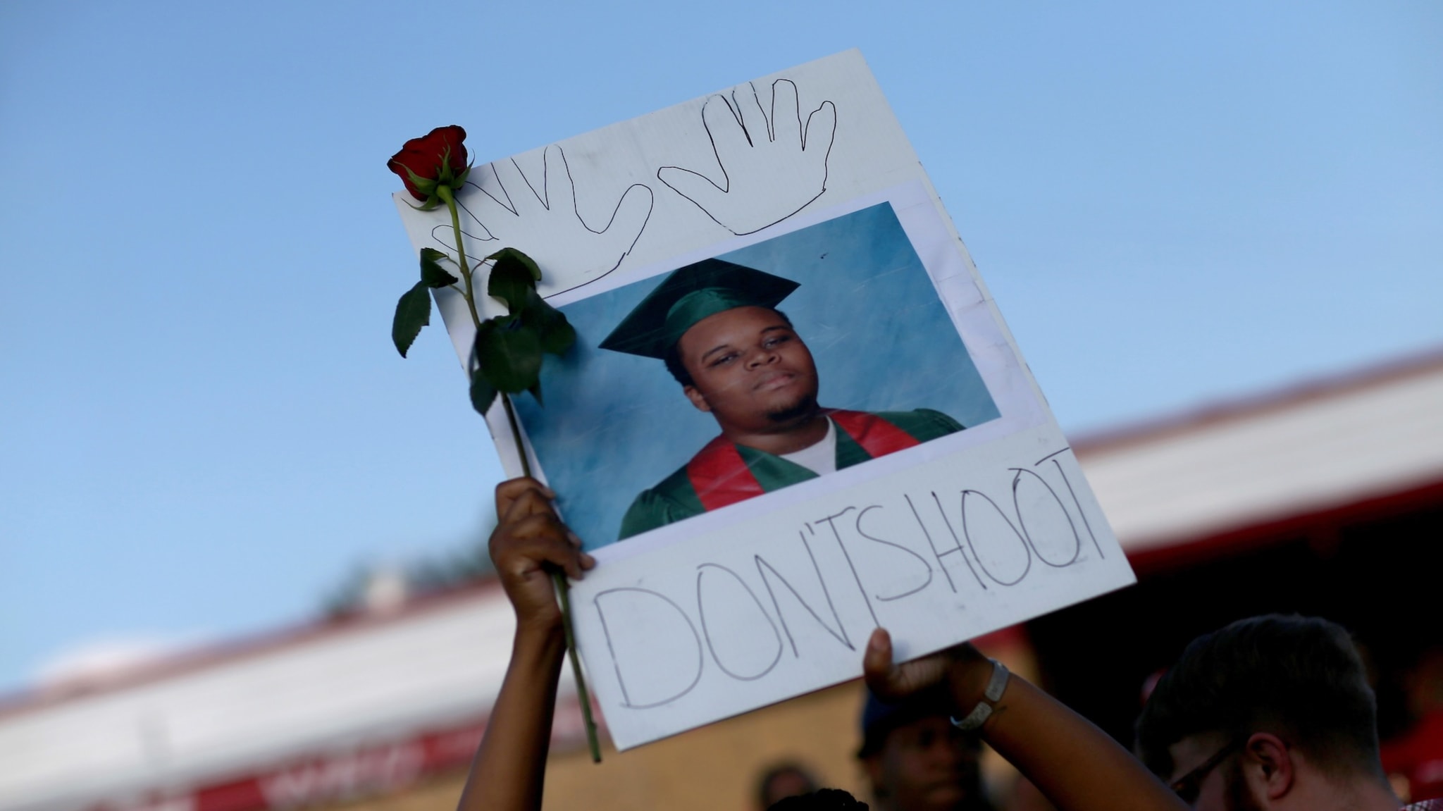 A protest sign with a photo of a young man in a graduation cap and gown, a rose, and the words "DON'T SHOOT" with hand outlines.