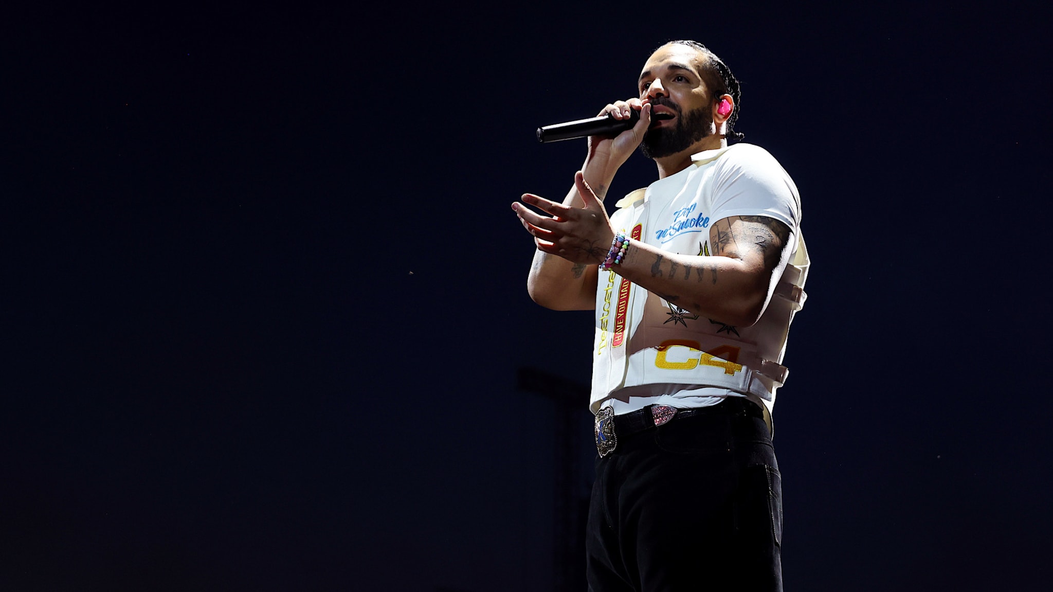 A man with tattoos and a beard performs on stage, holding a microphone, wearing a white t-shirt and black pants against a dark background.