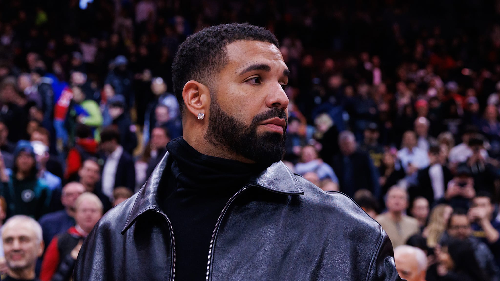 Drake, wearing a black leather jacket, stands in a crowd at an event.