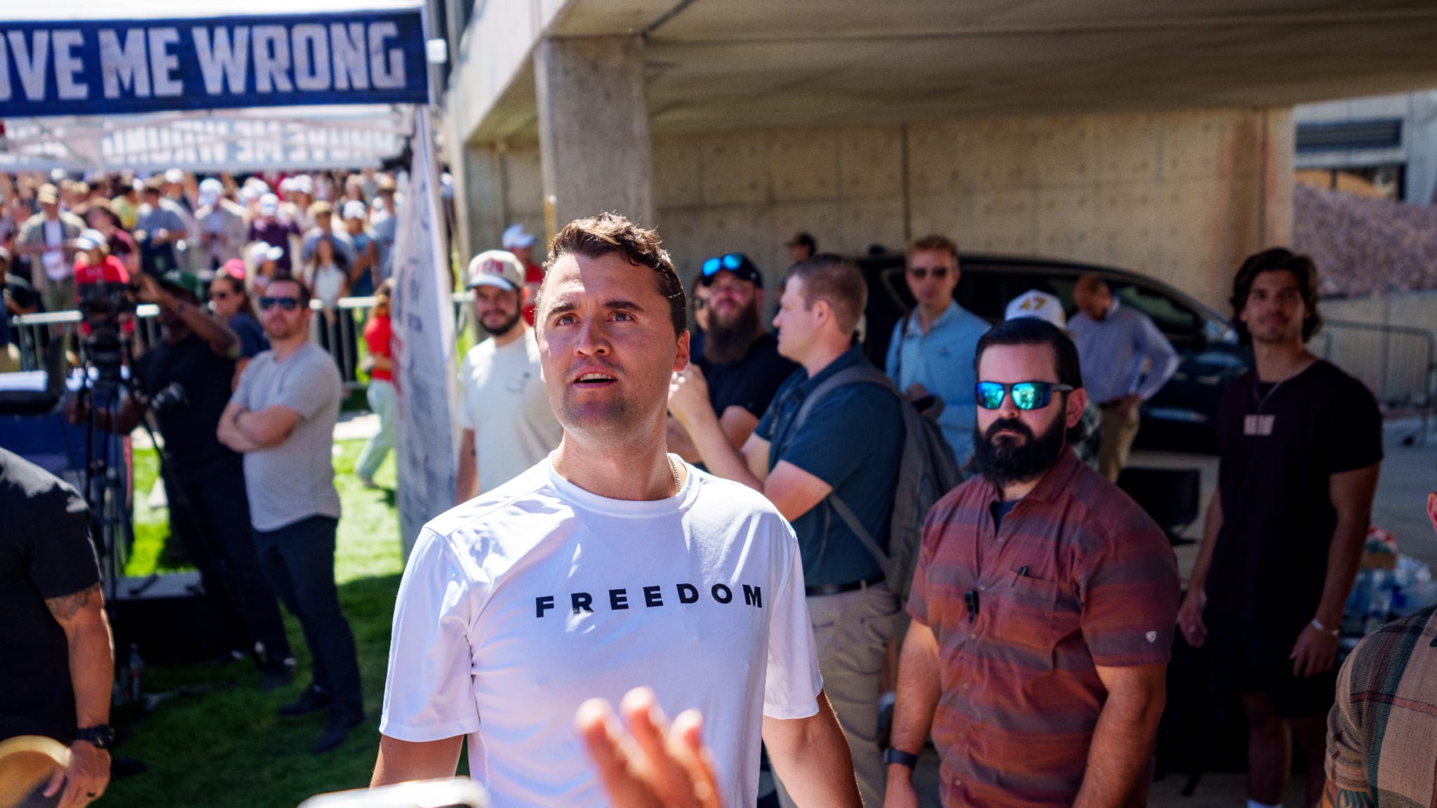 Charlie Kirk in a "FREEDOM" shirt stands among a crowd outdoors. People are gathered around, some wearing sunglasses. A banner reads "CHANGE MY MIND."