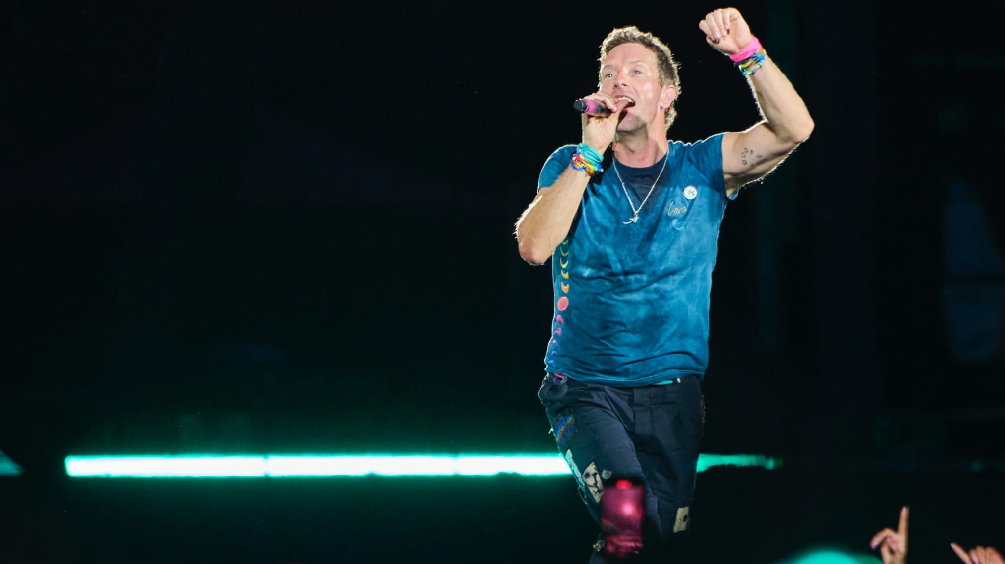 Chris Martin performing energetically on stage, wearing a blue shirt and colorful wristbands, with a microphone in hand.