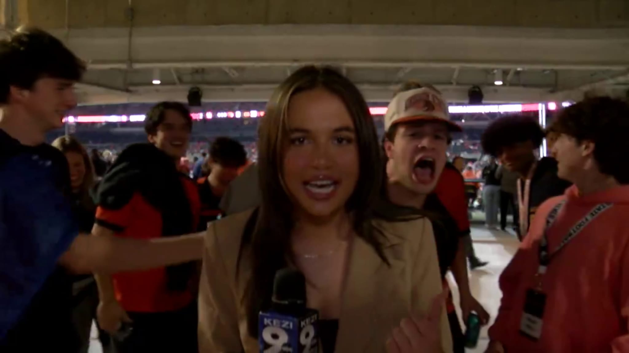 A reporter holding a KEZI 9 microphone is surrounded by excited people in a stadium. One person is shouting behind her.