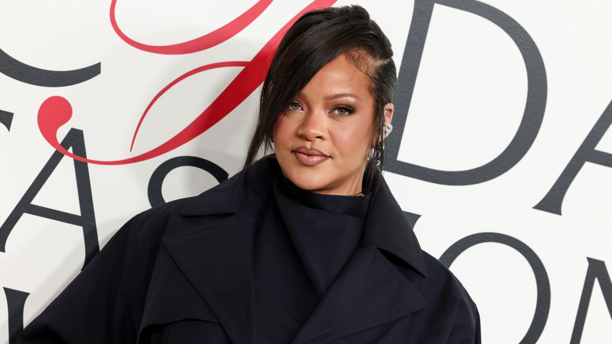 Rihanna at a fashion event, wearing a dark outfit with a side-swept hairstyle, standing in front of a stylized background.