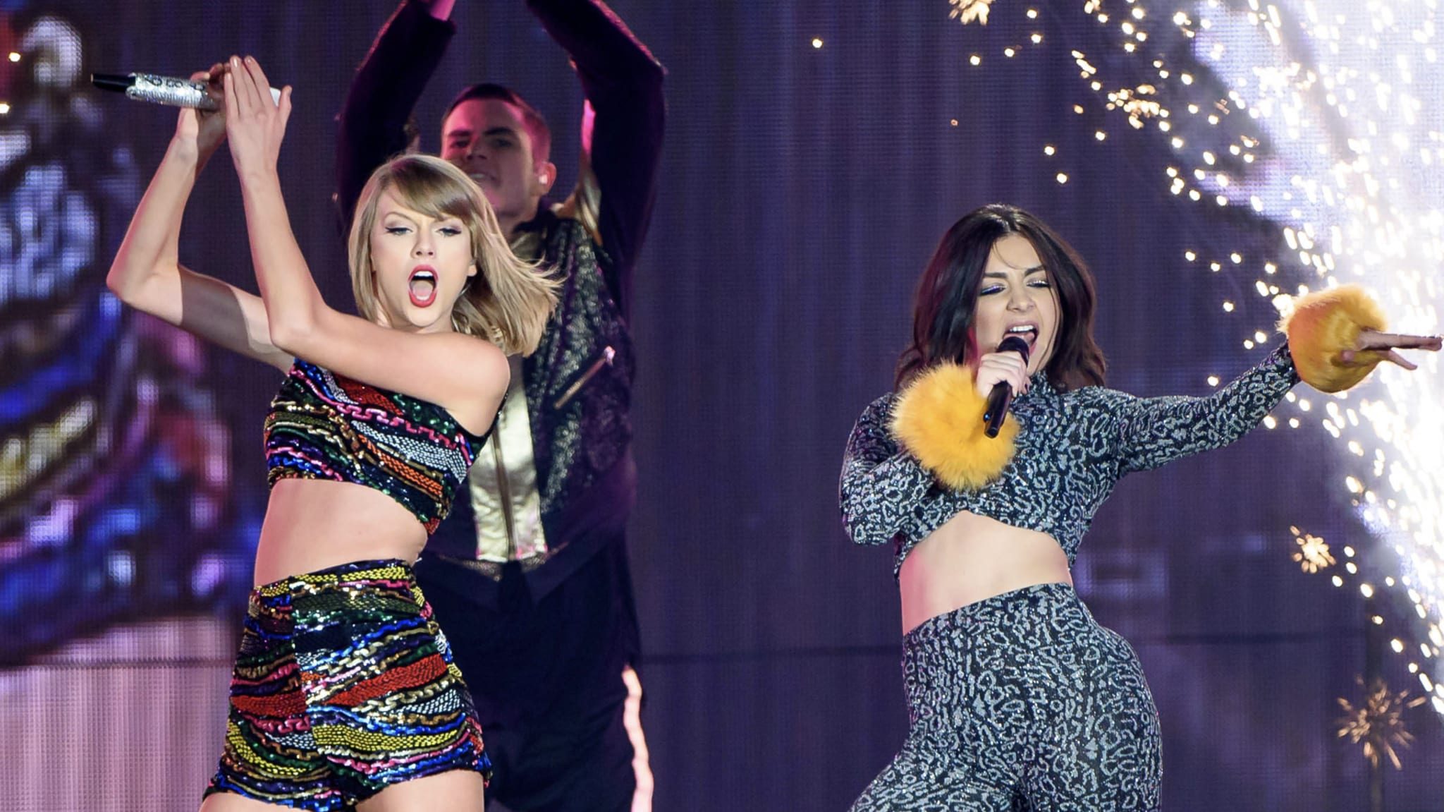 Taylor Swift and Chari xcx performing on stage, Swift in a colorful outfit, and Lorde in a leopard print ensemble with yellow cuffs.