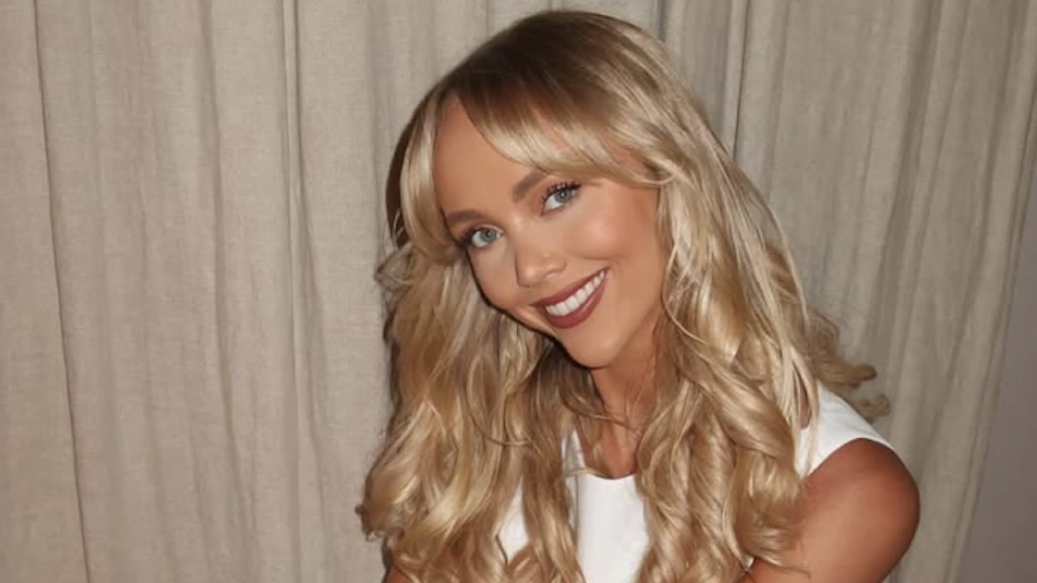 A woman with long blonde hair and a white top smiling in front of a curtain background.