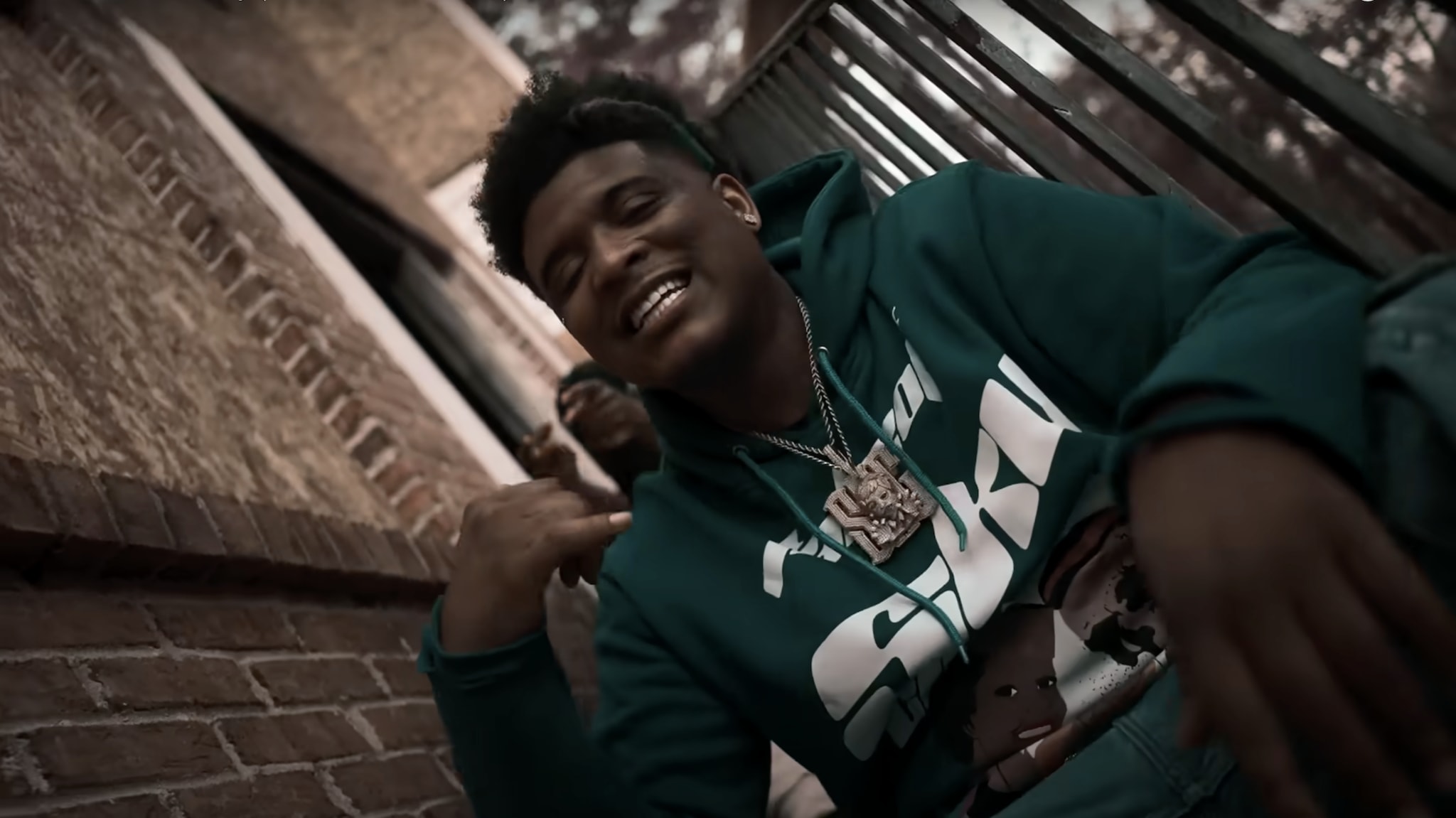 T-Hood in a green hoodie sits on steps, smiling and gesturing with their hand.