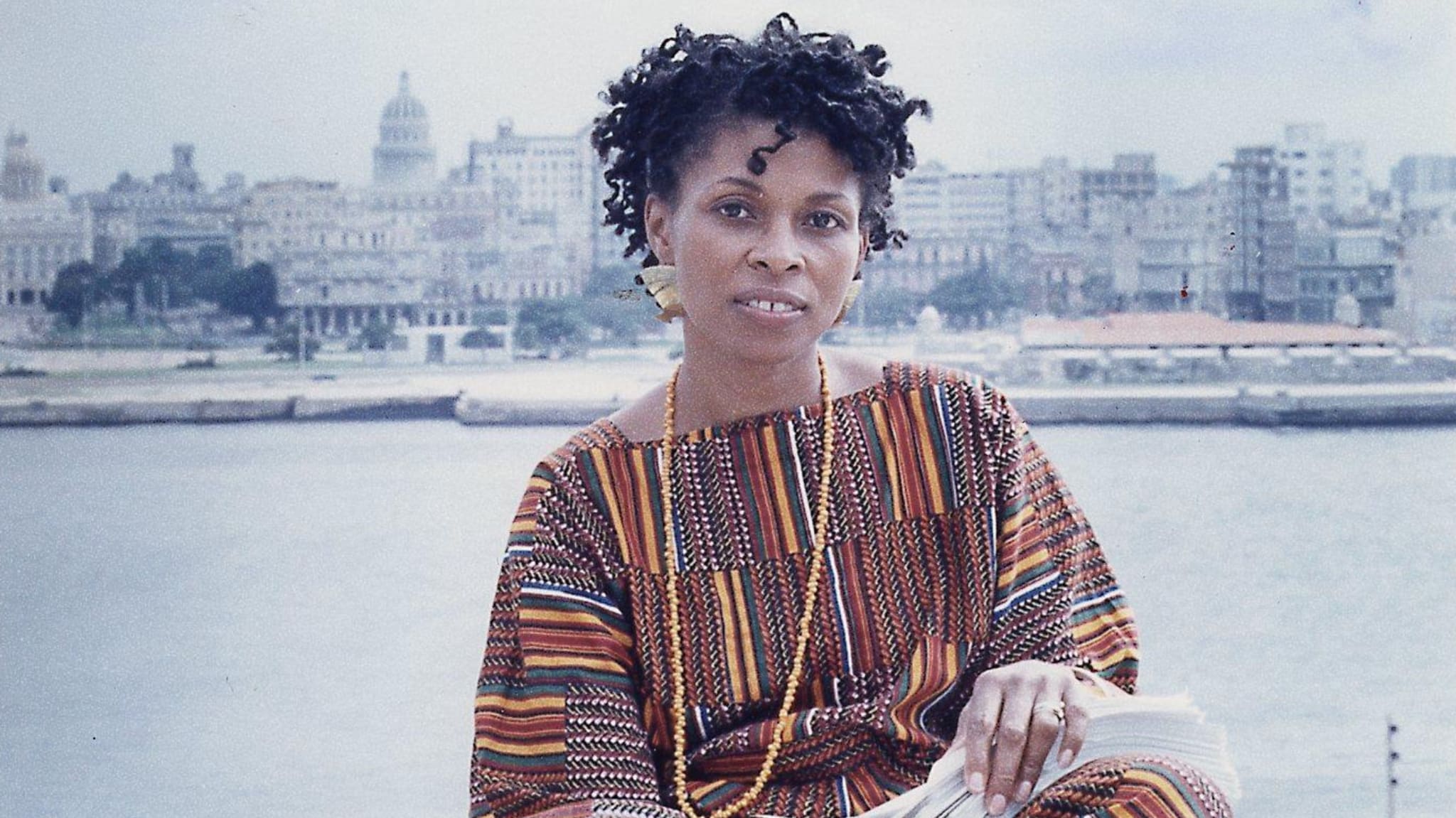 Assata Shakur in a colorful striped outfit sits by the water with a cityscape in the background. Her hair is styled in short curls.