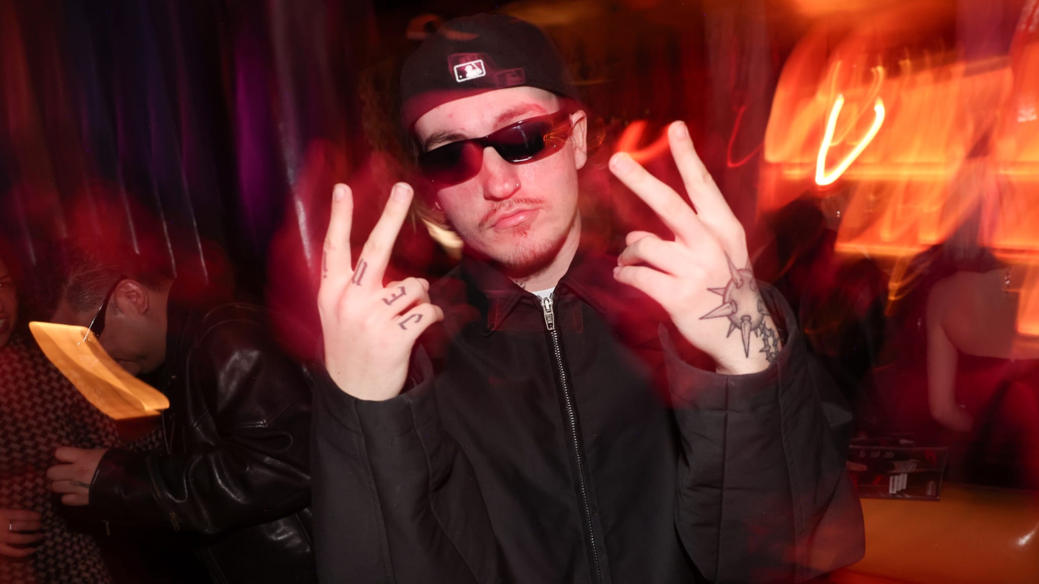 Tommy Richman in a dark jacket, cap, and sunglasses poses with peace signs in a dimly lit room with red and orange lights.