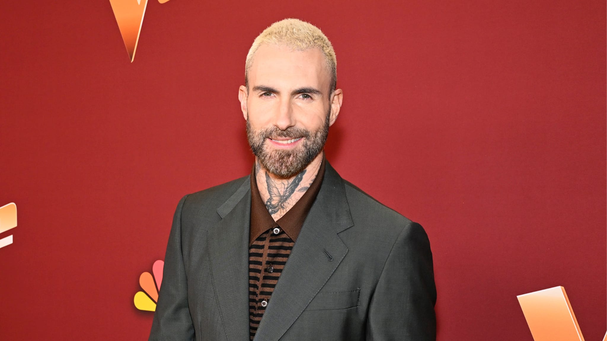 Adam Levine with blonde hair and a beard, wearing a dark suit against a red background.