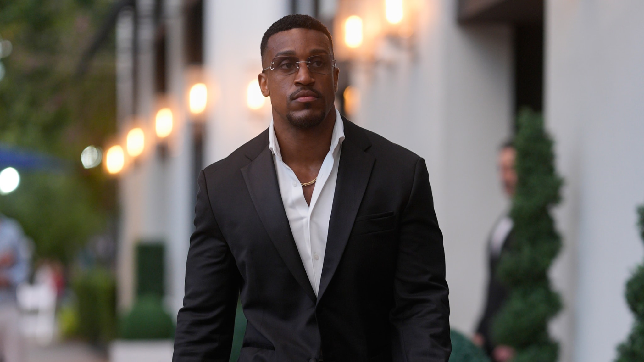 Fitness influencer Ashton Hall in a black suit and glasses walks on a street lined with lights and greenery.