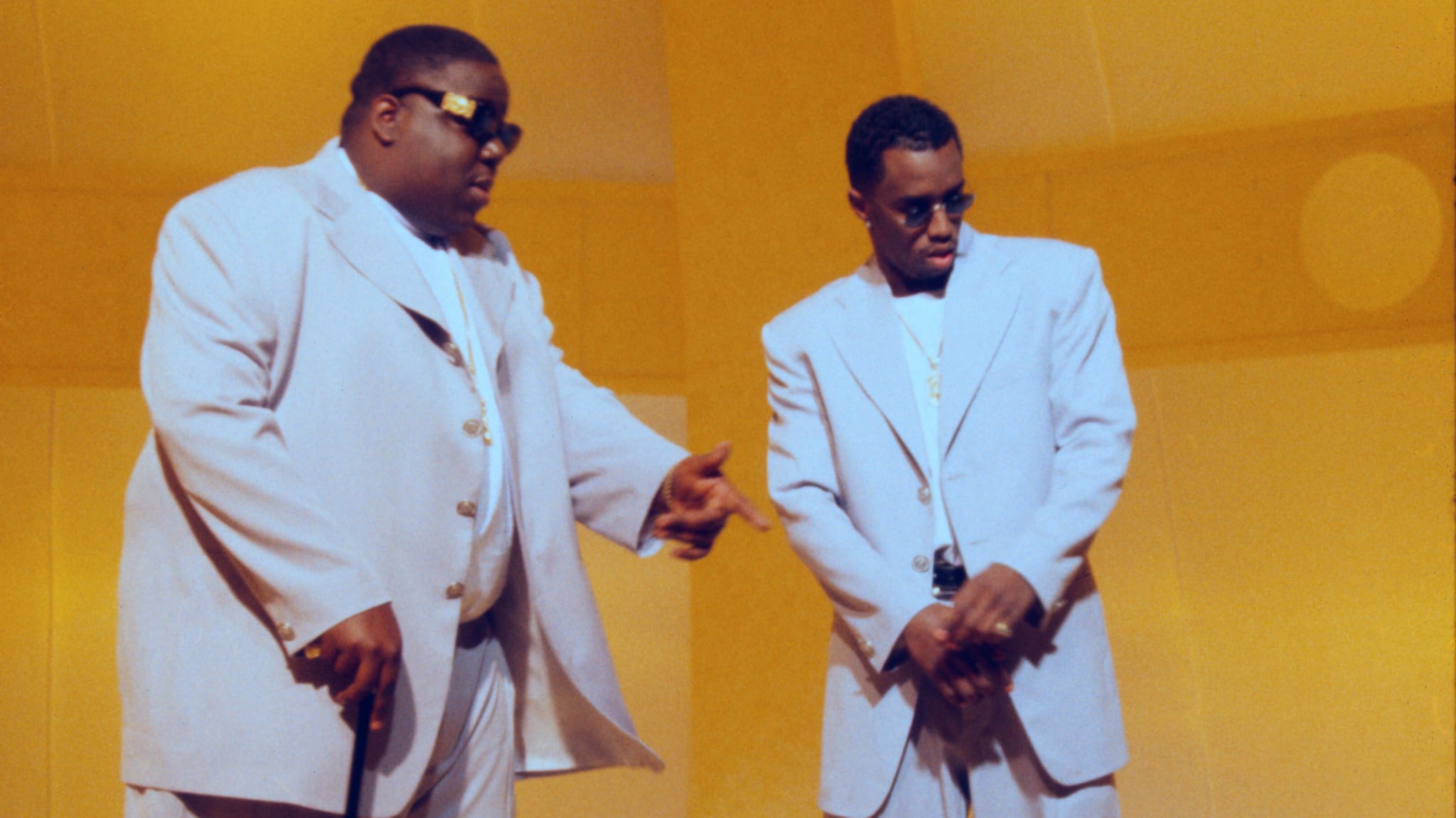 The image shows The Notorious B.I.G. and Sean "Diddy" Combs in matching light suits, standing in a yellow-toned setting.