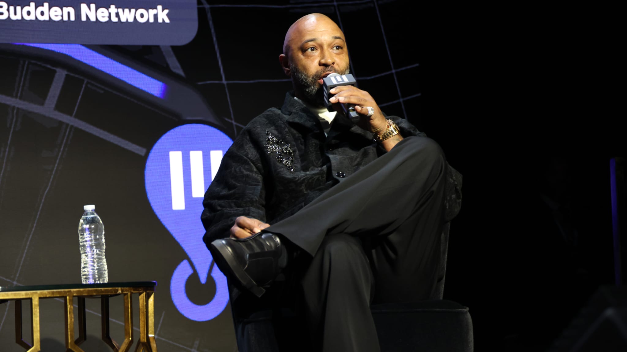 Joe Budden sitting on stage, speaking into a microphone, with a water bottle on a table nearby.