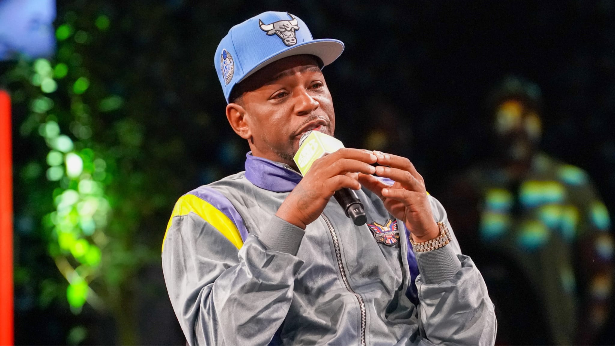 A man in a blue Chicago Bulls cap and gray jacket speaks into a microphone on stage.