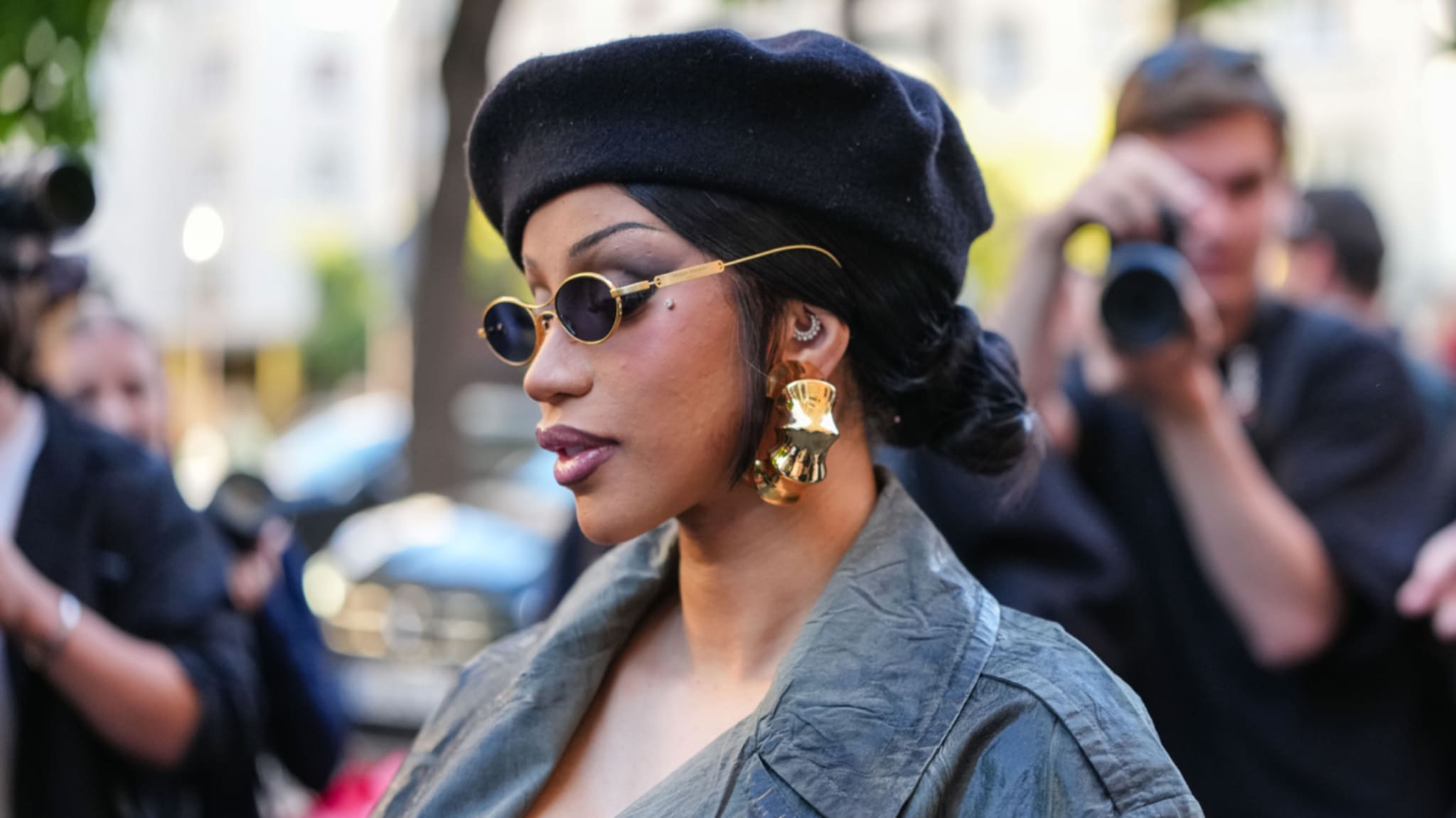 Cardi B in a black beret and sunglasses, wearing large gold earrings, is surrounded by photographers.