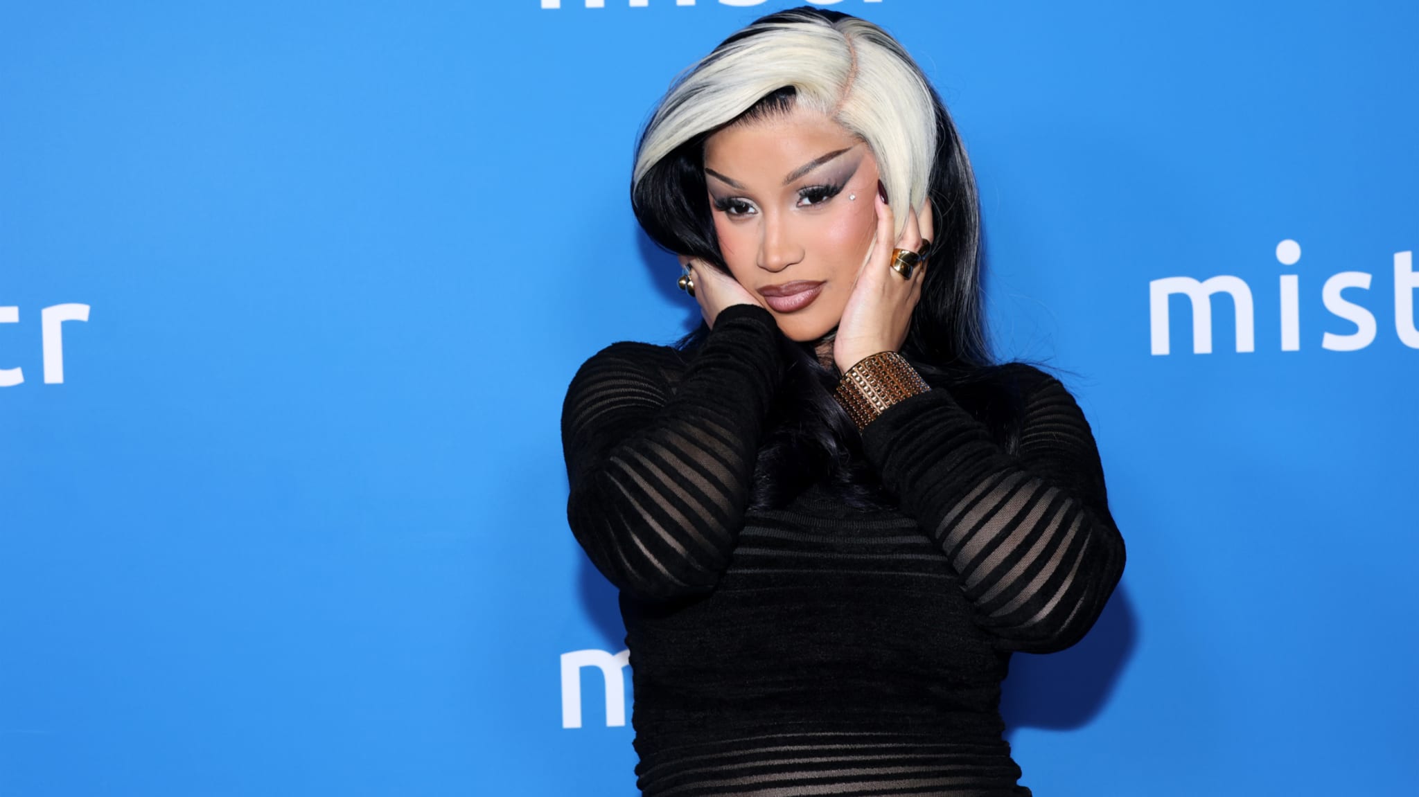 Cardi B with black and white hair poses in front of a blue background with "mistr" text, wearing a black outfit and gold accessories.