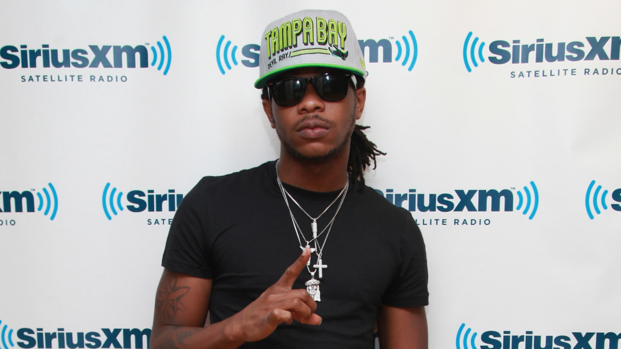 Rapper Cash Out wearing a "Tampa Bay" cap, sunglasses, and necklaces, posing in front of a backdrop.