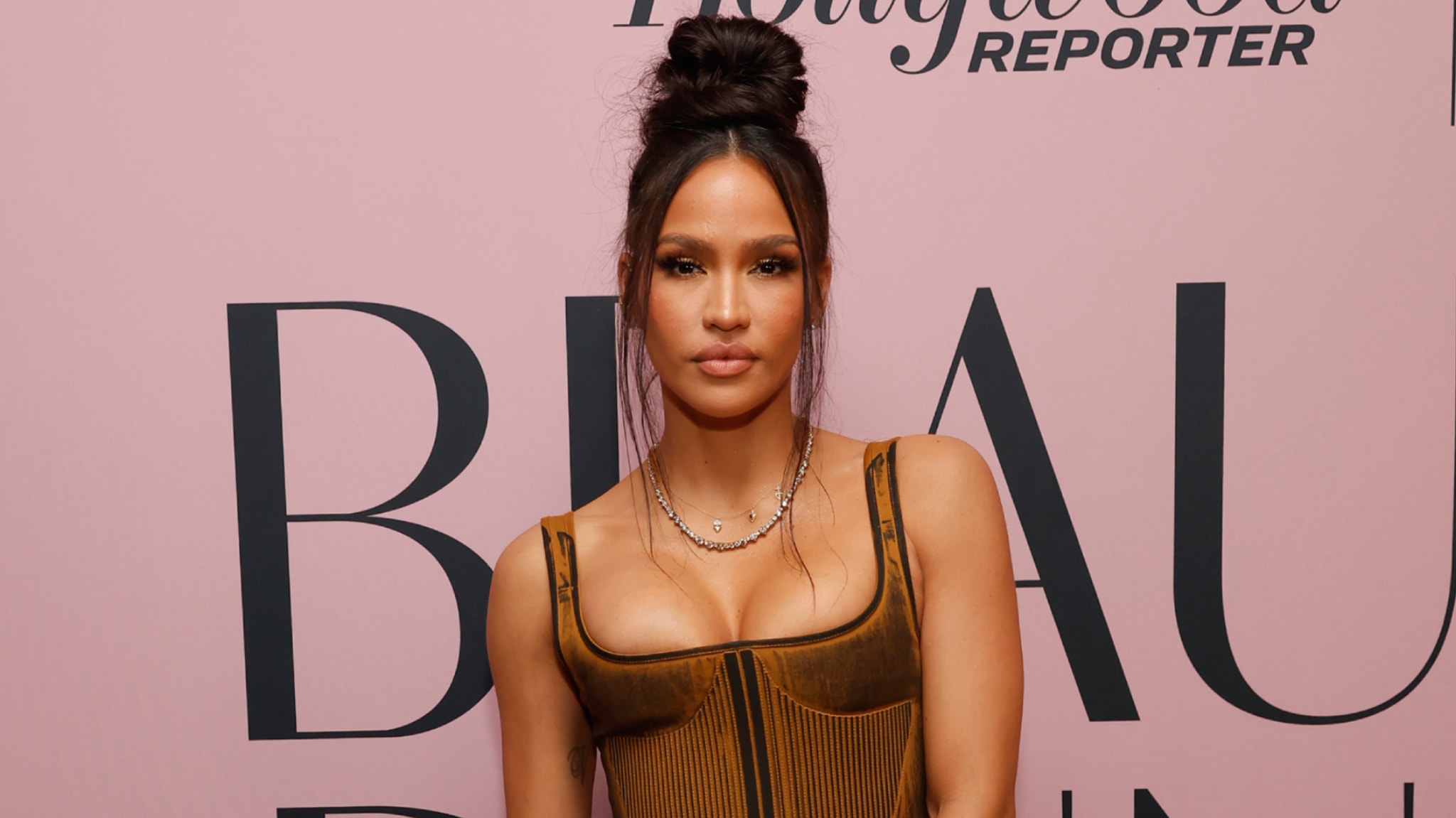 Cassie Ventura circa 2003 with long hair in a high bun, wearing a brown corset top, stands in front of a pink backdrop with text.