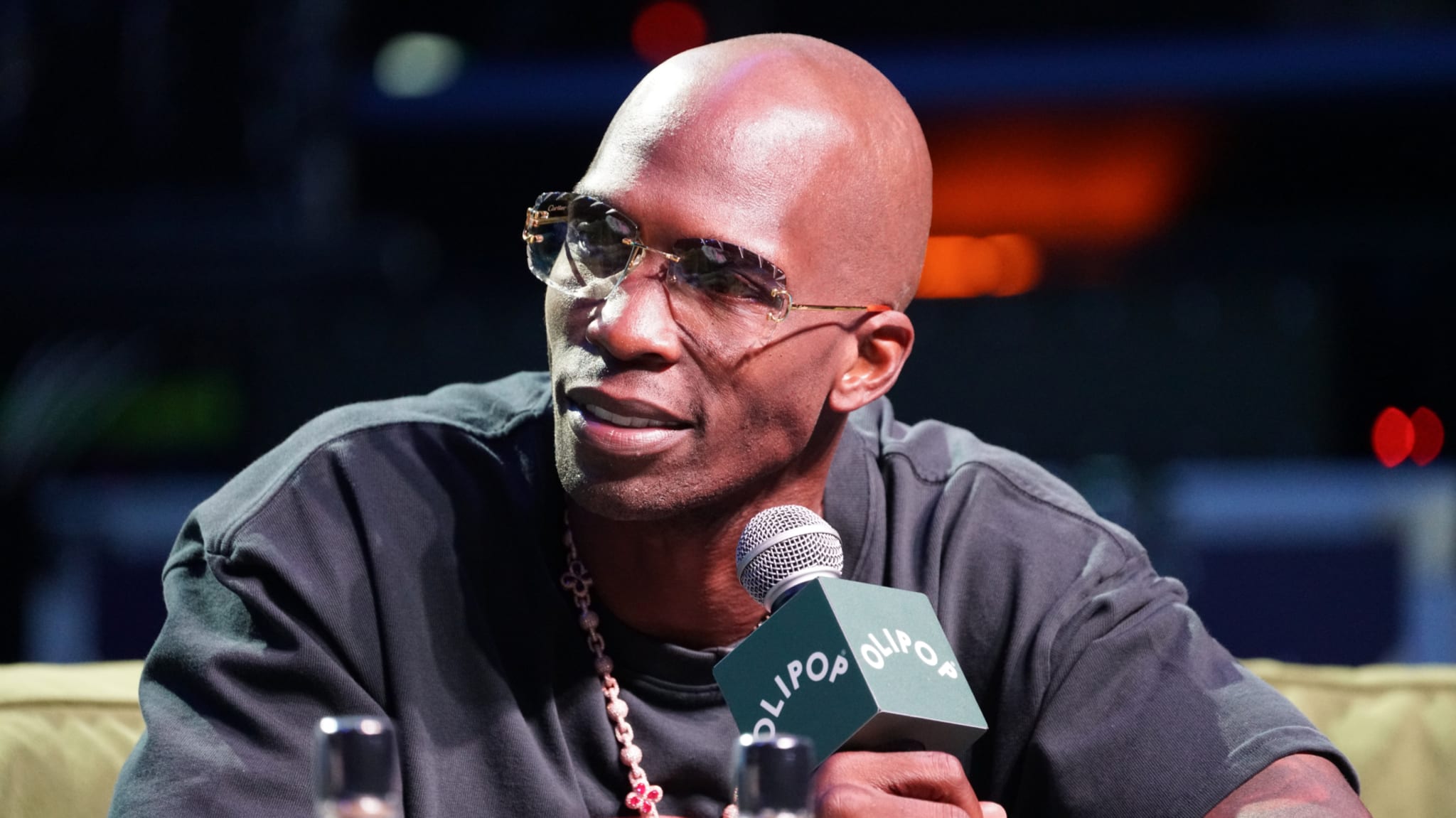 Chad Ochocinco Johnson wearing sunglasses and a dark shirt speaks into a microphone on stage.