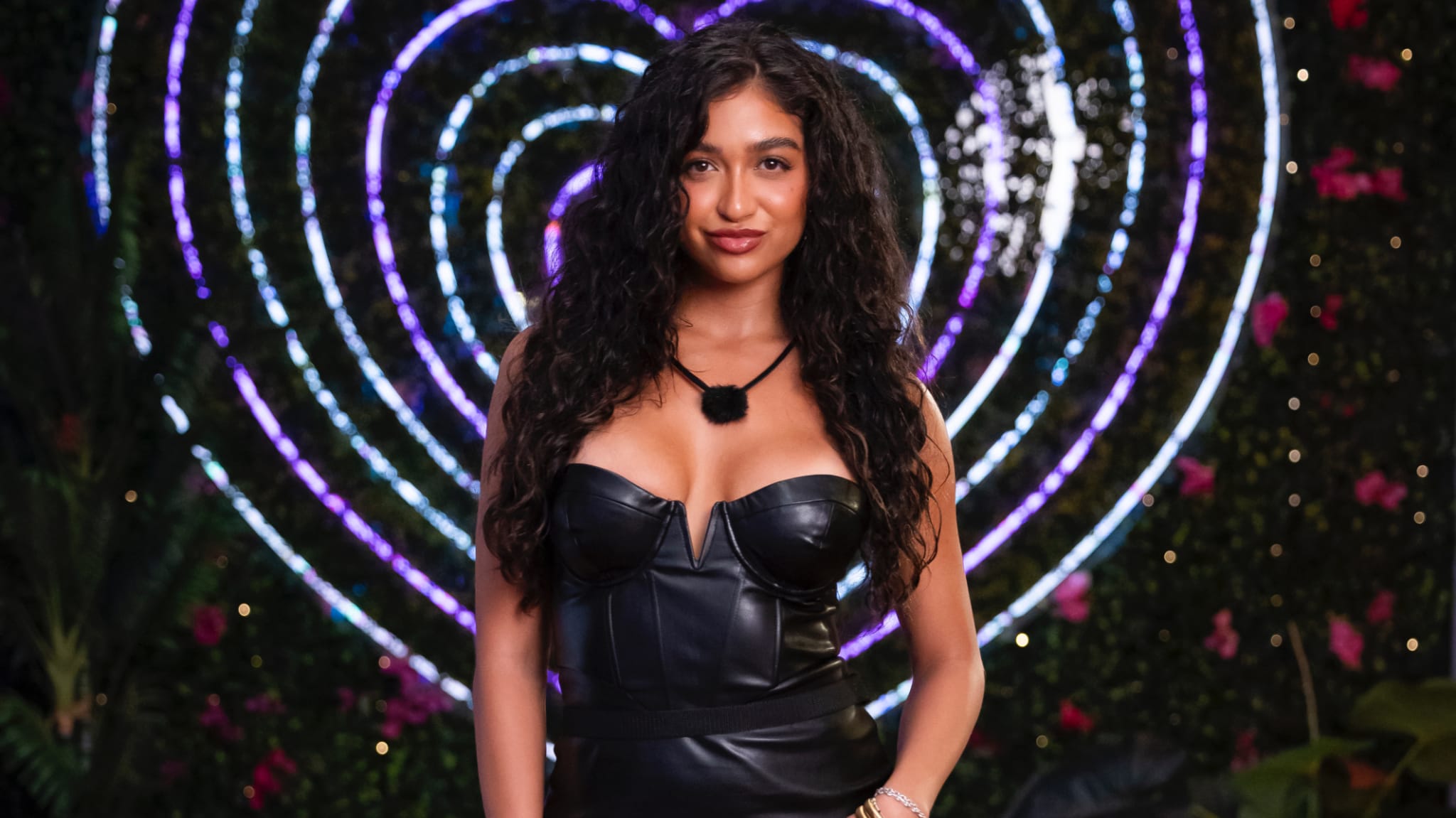 Cierra Ortega in a black leather outfit stands in front of a heart-shaped neon light on a dark, leafy background.
