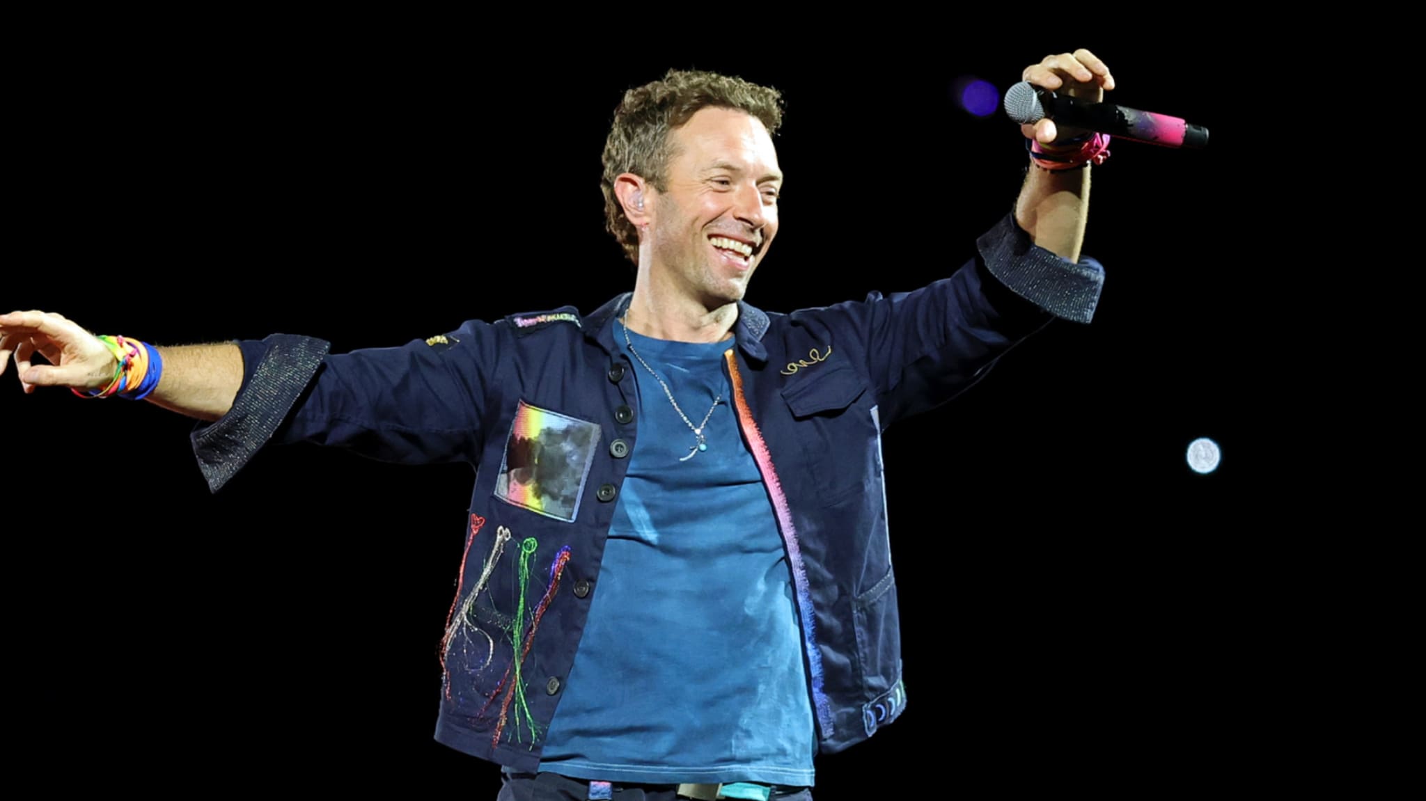 Chris Martin performing on stage, wearing a colorful jacket and holding a microphone, smiling with arms outstretched.