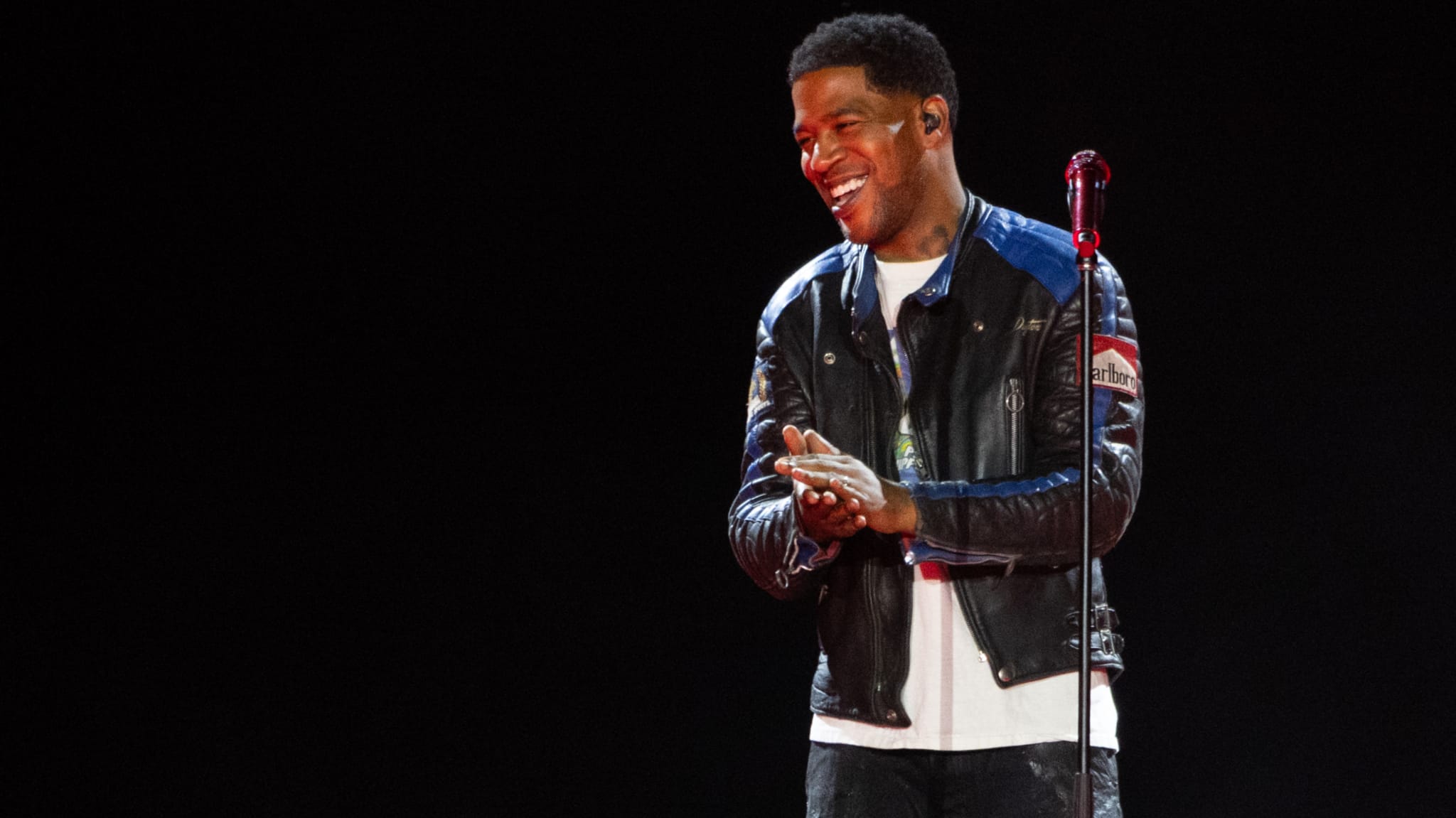 Kid Cudi performing on stage, wearing a leather jacket, smiling and clapping in front of a microphone.