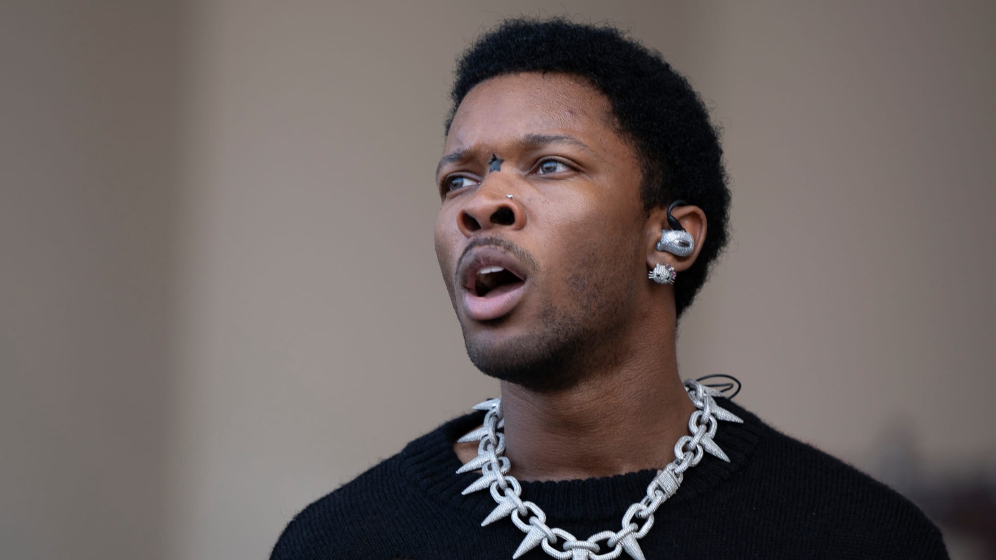 Singer D4vd with short hair, wearing a chain necklace and multiple earrings, looks upwards with an expressive face.