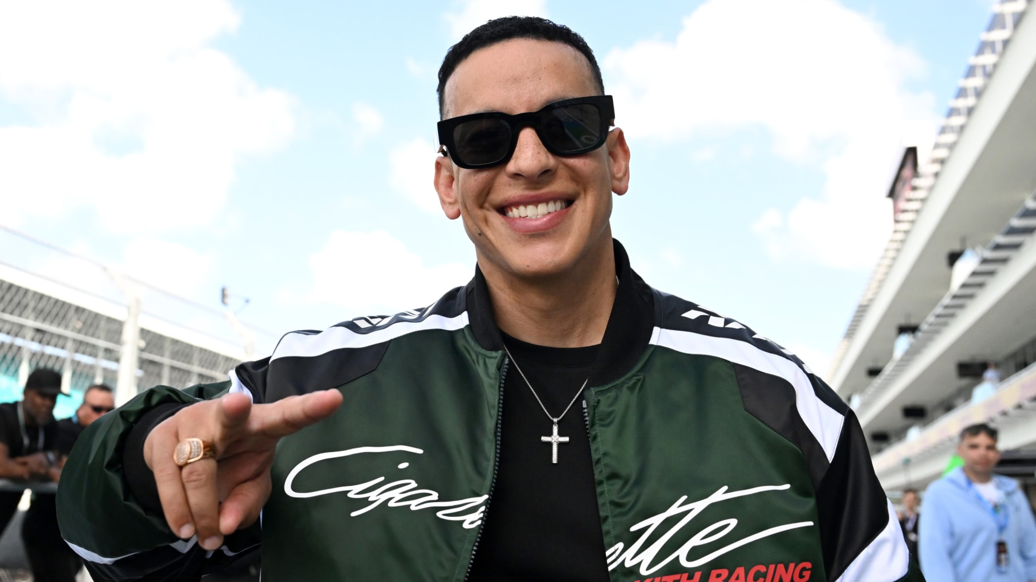 Daddy Yankee smiling, wearing sunglasses and a racing jacket, making a peace sign gesture outdoors.