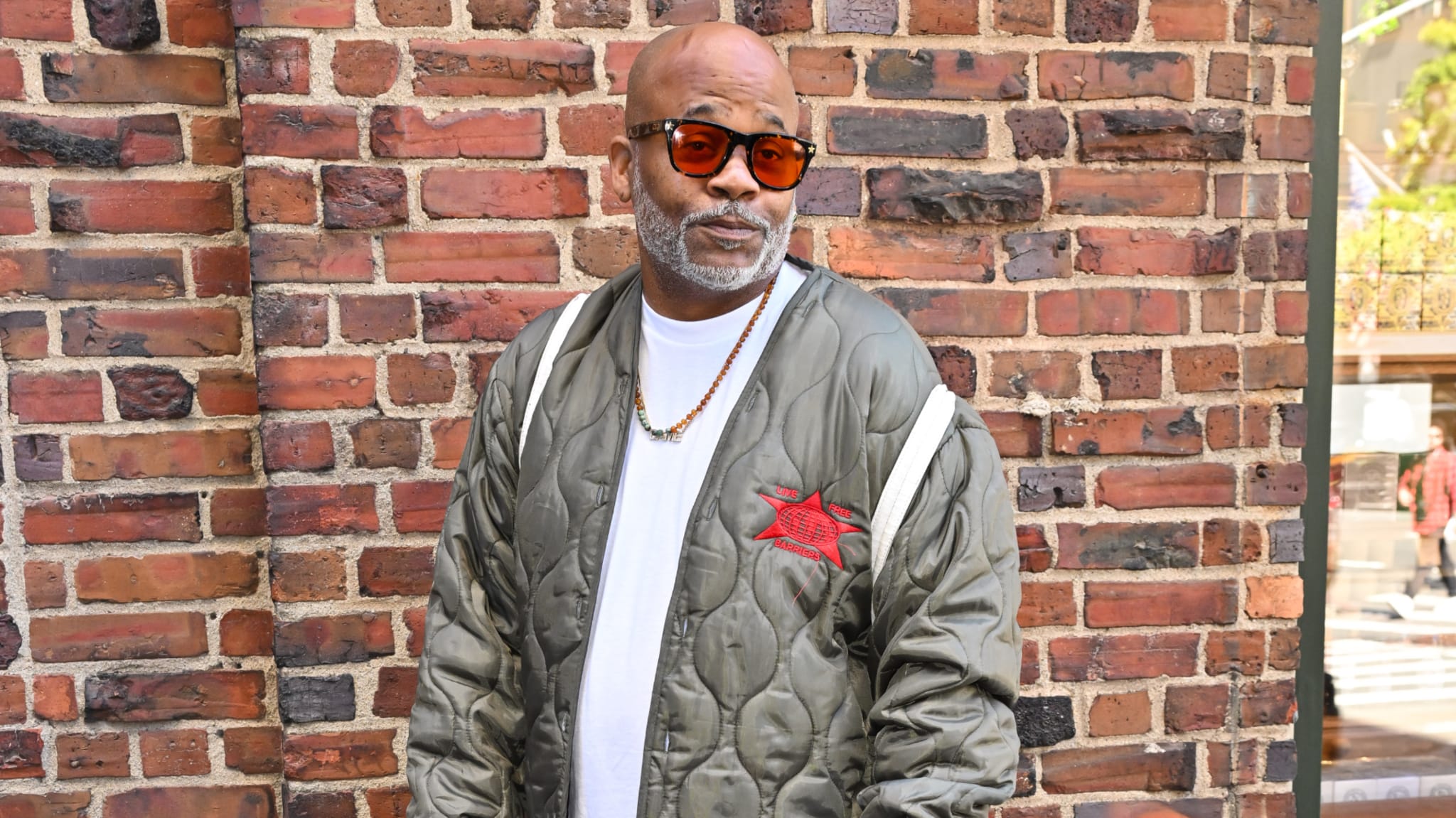 Dame Dash in sunglasses and a green quilted jacket stands against a brick wall, wearing a white shirt and necklace.